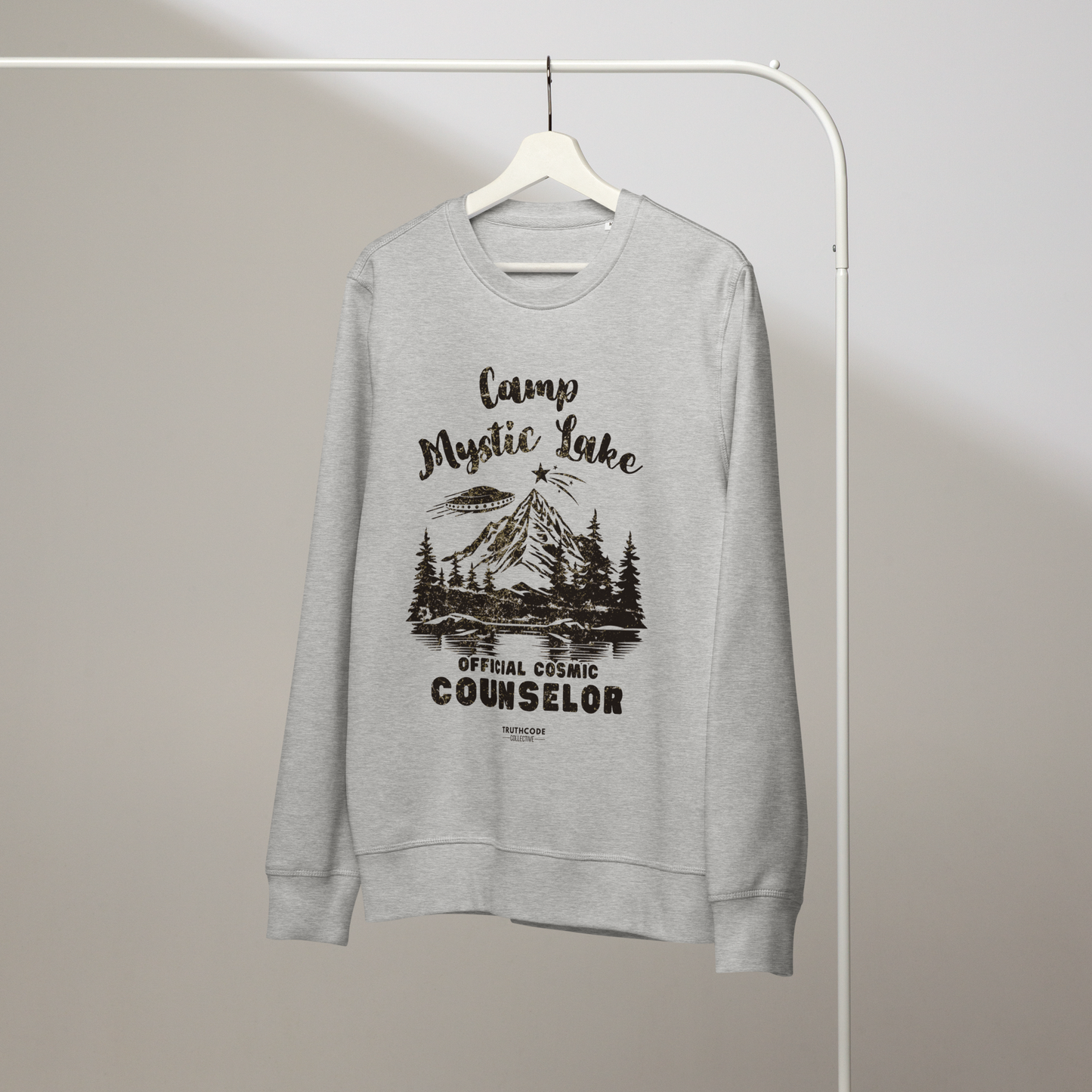 Gray organic sweatshirt with mountain scene and spaceship graphic design and text 'Camp Mystic Lake, official cosmic counselor' on a white hanger with a gray wall background