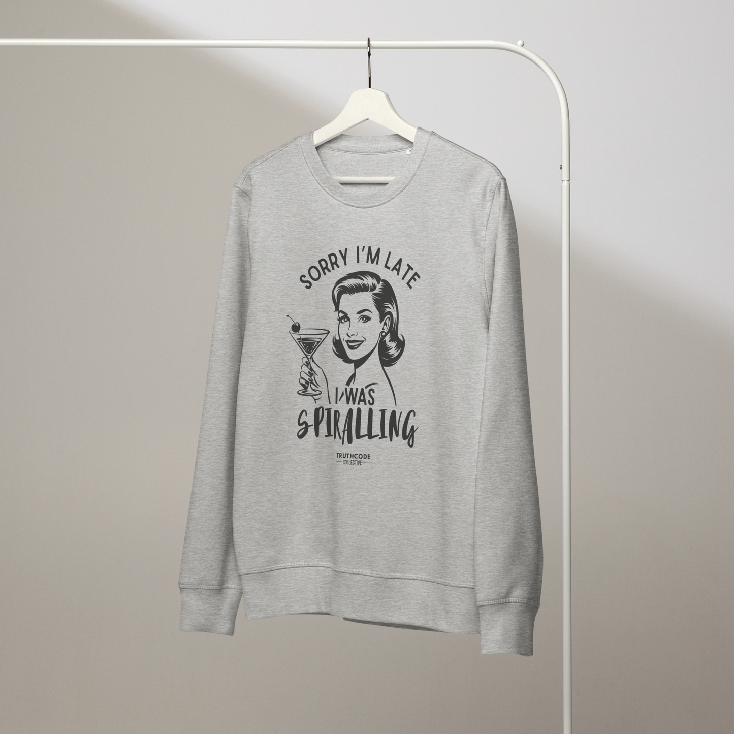 Gray organic sweatshirt with vintage graphic design of woman holding a drink and text 'Sorry I'm late I was spiraling' on a white hanger with a gray wall background