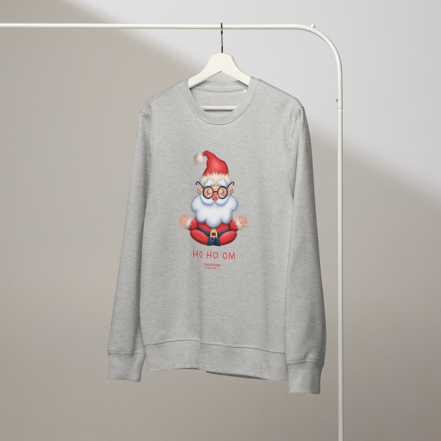 Gray organic sweatshirt with a cartoon Santa Claus meditating design and text 'HO HO OM' on a hanger with a gray wall background