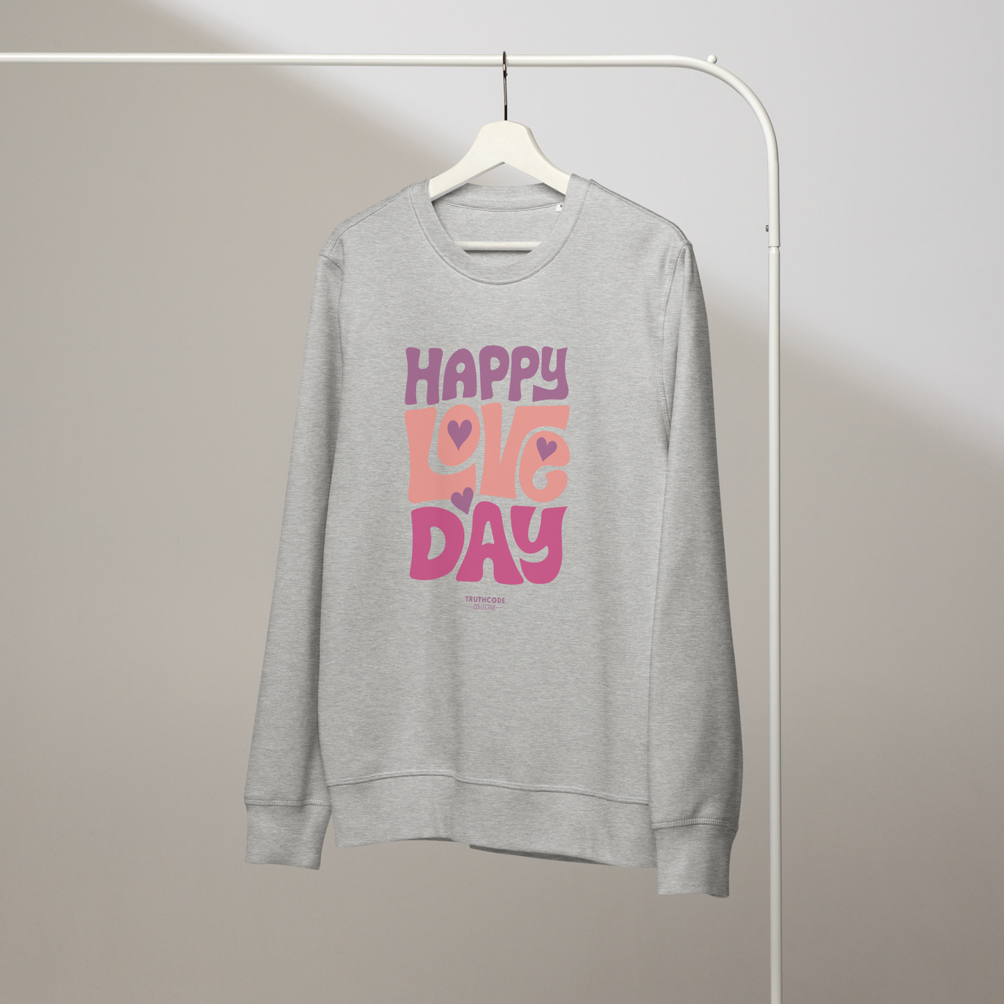 Gray organic sweatshirt with 'Happy Love Day' text in pink and purple on a hanger with a gray wall background