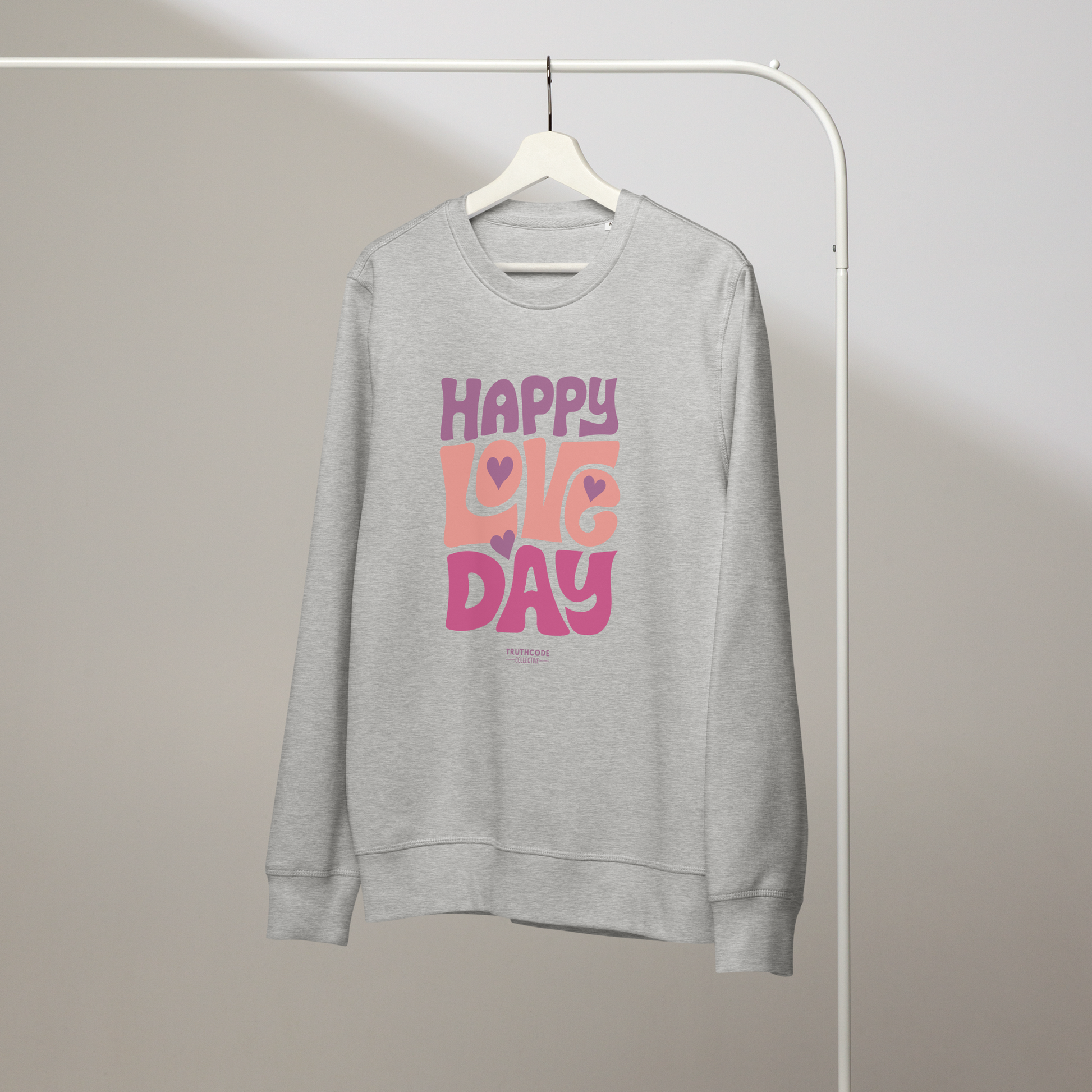 Gray organic sweatshirt with 'Happy Love Day' text in pink and purple on a hanger with a gray wall background