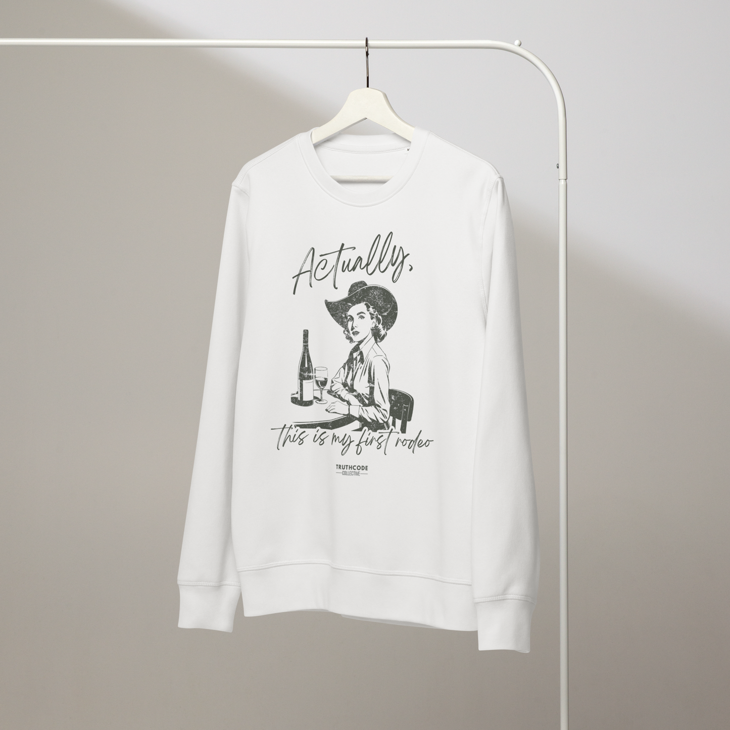 White organic sweatshirt with vintage graphic of a woman in a cowboy hat and text 'Actually, this is my first rodeo' on a white hanger with a gray wall background