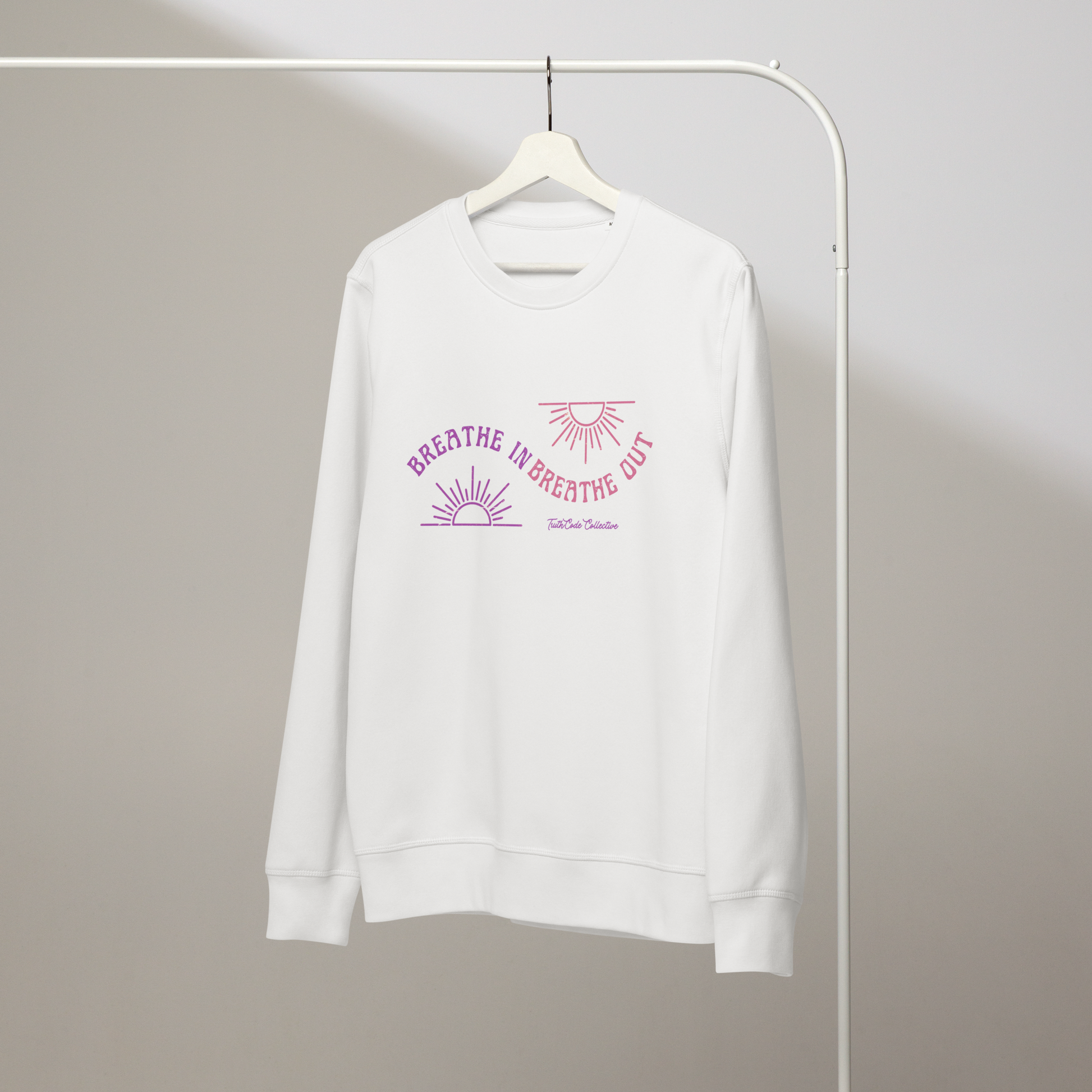 White organic sweatshirt with pink and purple sun design and text 'Breathe in breathe out' on a white hanger with a gray wall background