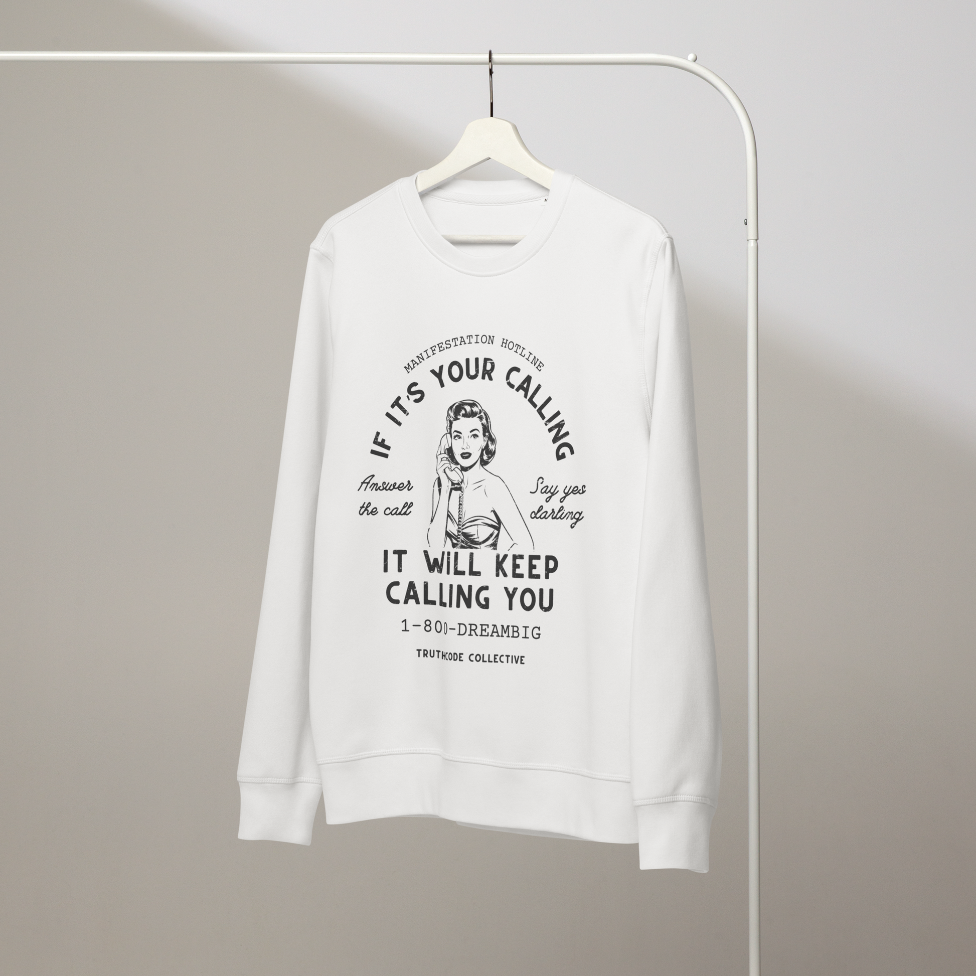 White organic sweatshirt with vintage-style graphic of a woman on the phone and text 'If it's your calling, it will keep calling you' and '1-800-DREAMBIG' on a white hanger with a gray wall background