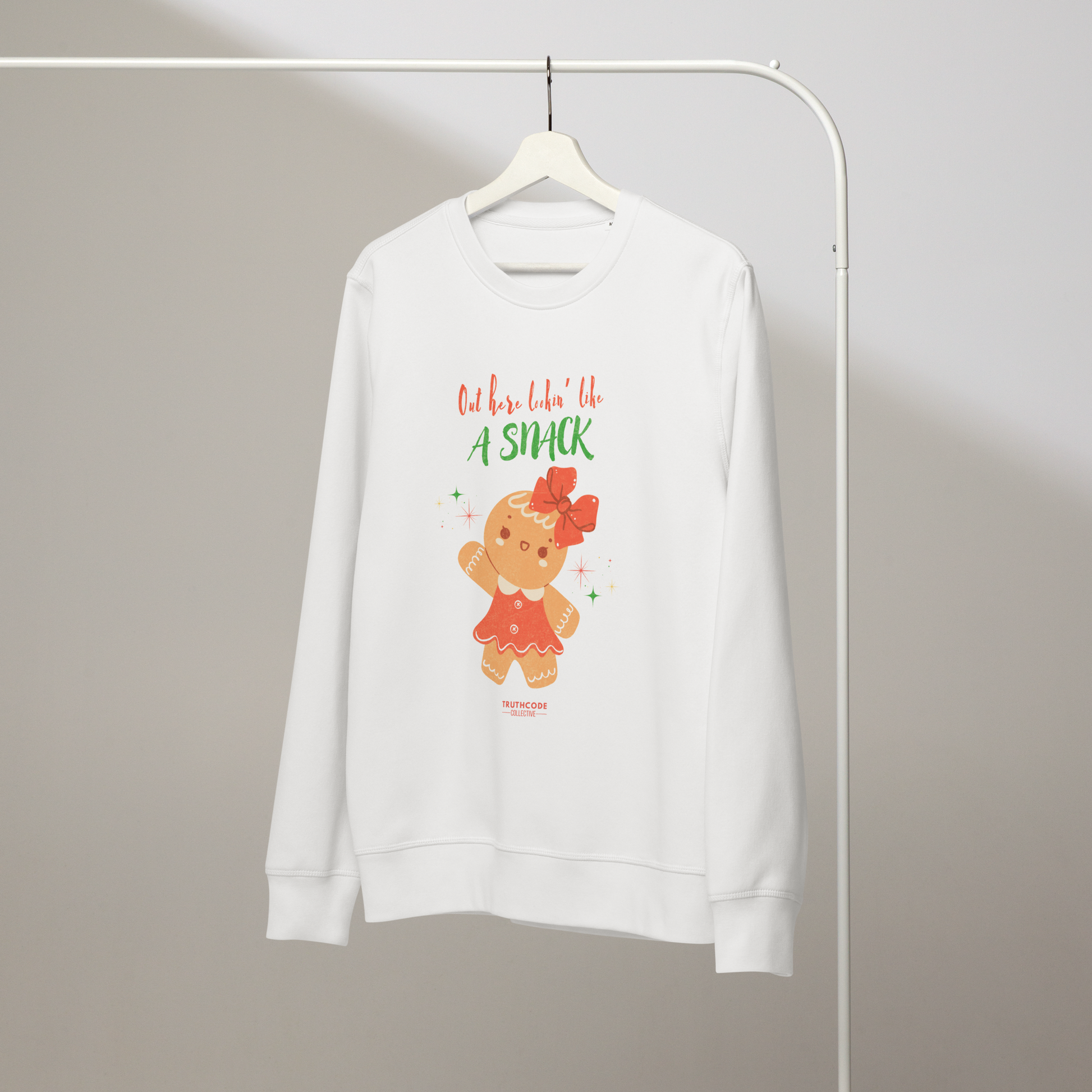 White organic sweatshirt with a gingerbread girl graphic and text 'Out here lookin' like a snack' on a hanger with a gray wall background