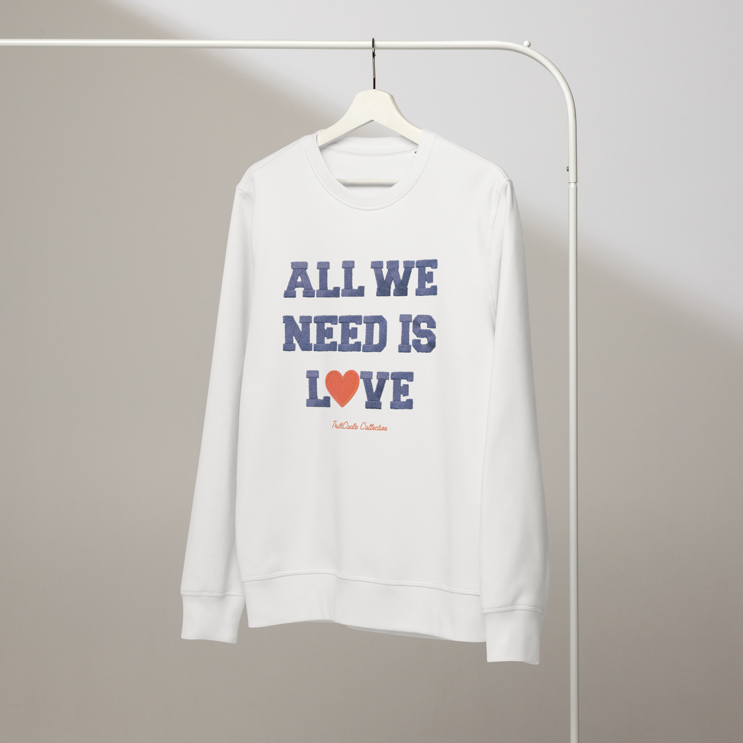 White organic sweatshirt with 'All We Need Is Love' text and heart graphic on a hanger with a gray wall background