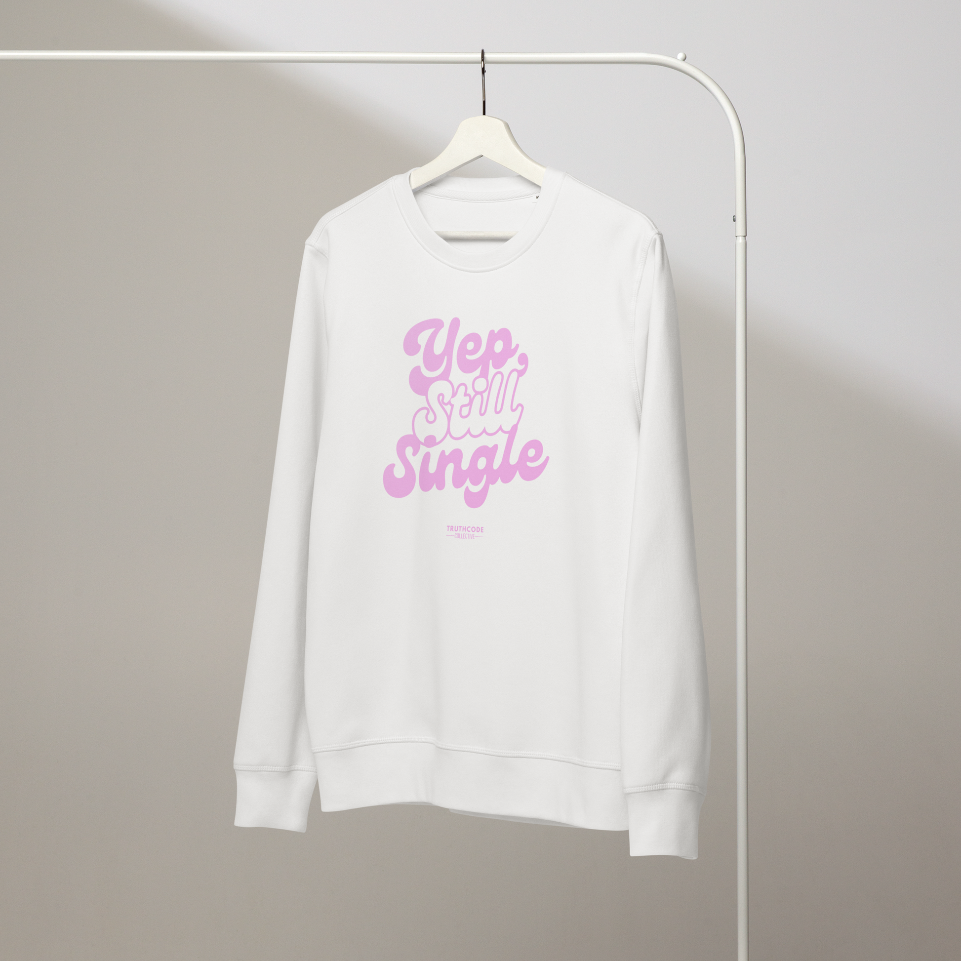 White organic sweatshirt with pink text 'Yep, still single', on a hanger with a gray wall background