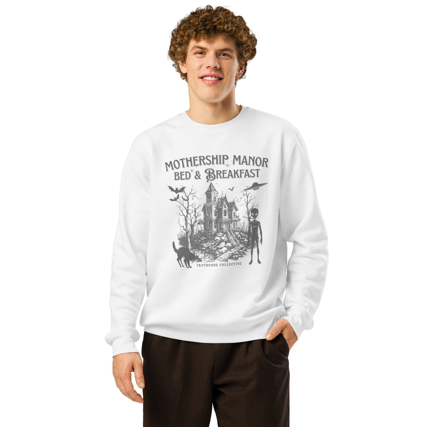 Model wearing beige organic sweatshirt with a graphic design of a haunted house, alien, and spaceship, with text 'Mothership Manor Bed & Breakfast' on a white background