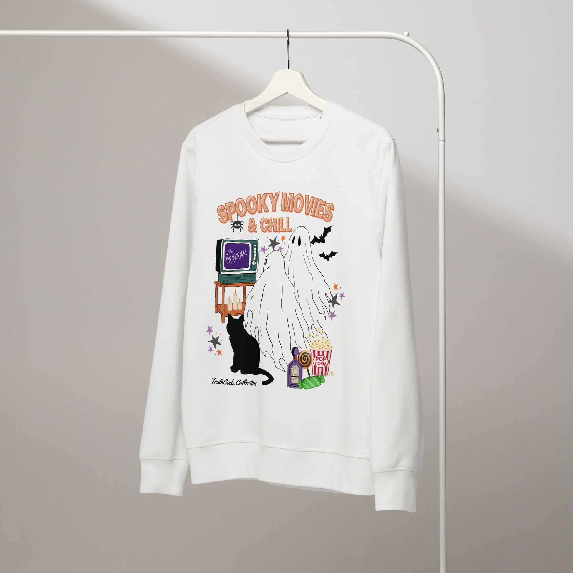 White organic sweatshirt with Halloween-themed design featuring a ghost, black cat, TV, and popcorn and text 'Spooky movies and chill' on a hanger with a gray wall background