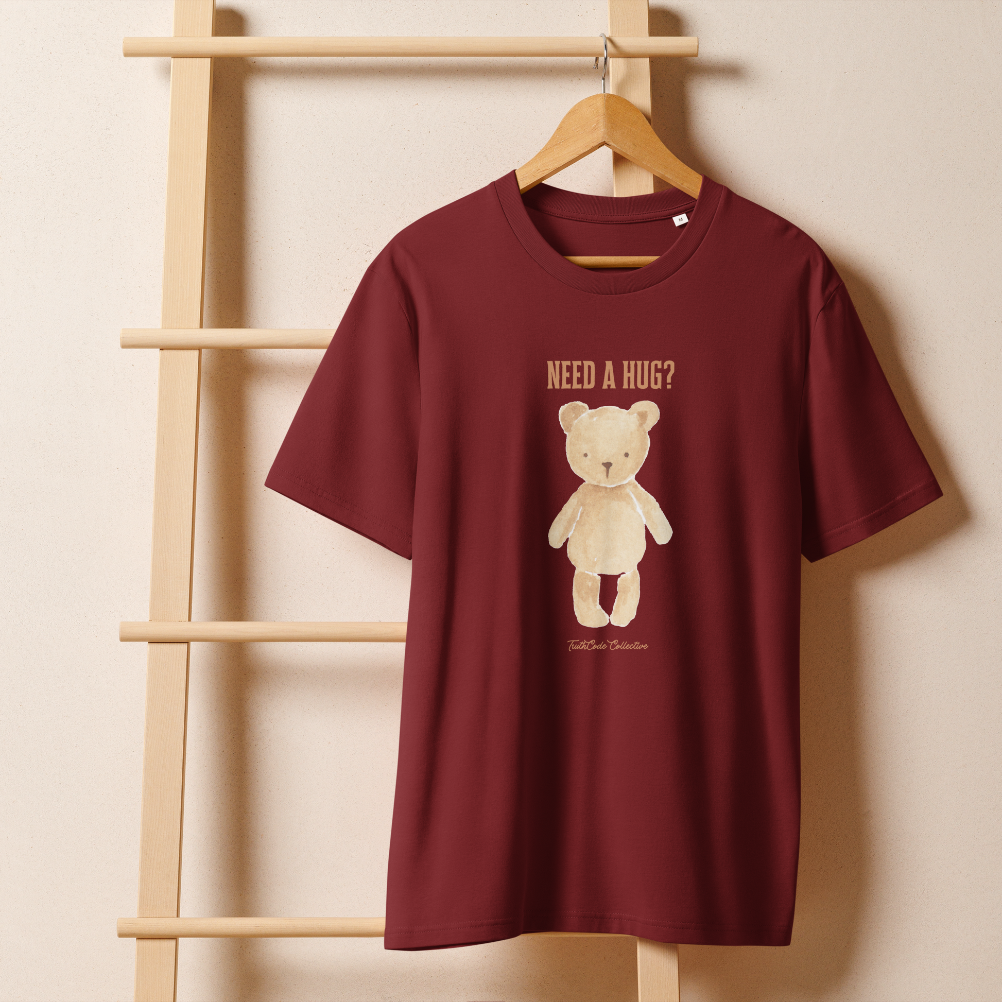 Burgundy organic t-shirt with a teddy bear graphic and text 'NEED A HUG?' on a wooden hanger with a beige wall background