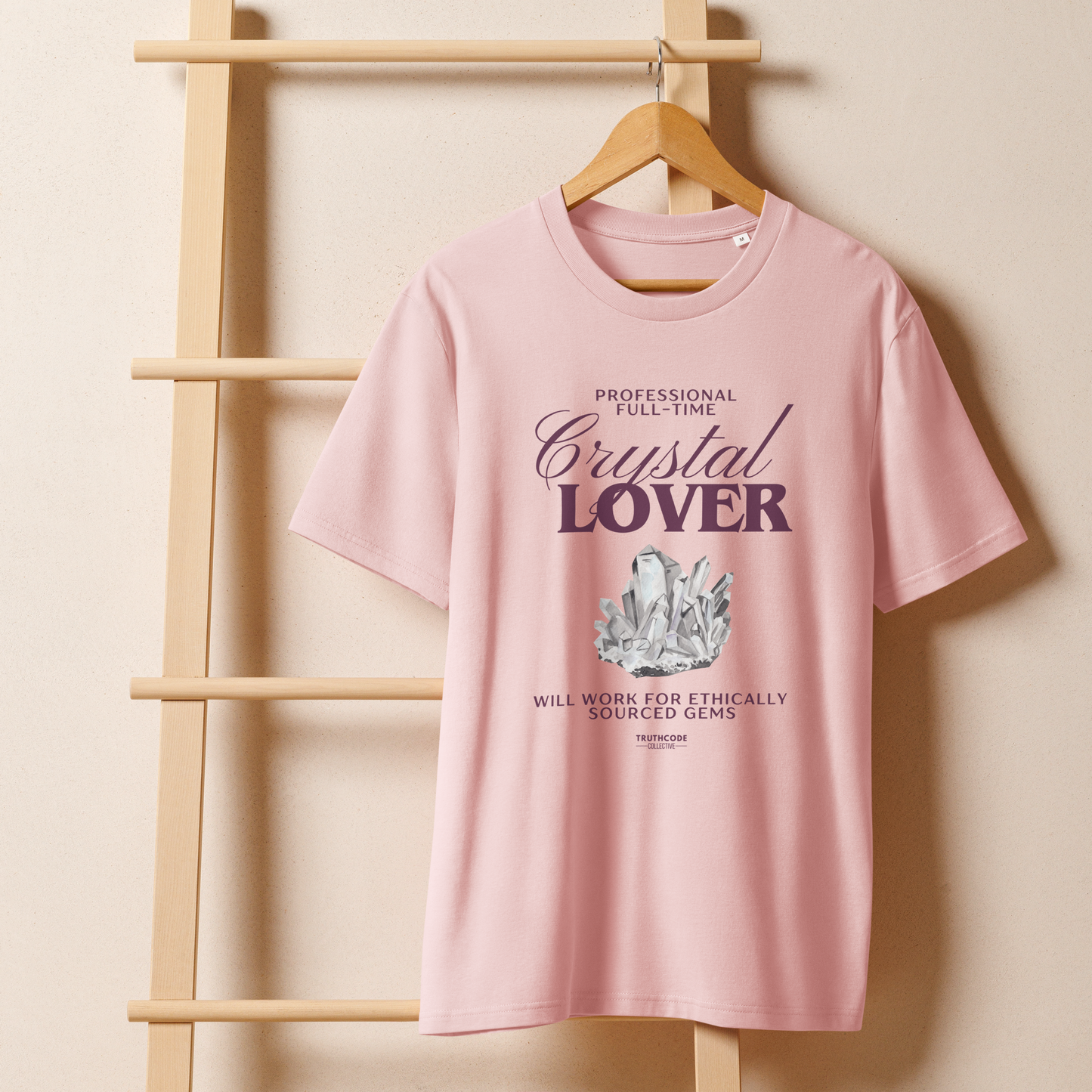 Pink organic t-shirt with crystal graphic and text saying 'Crystal Lover, will work for ethically sourced gems' hanging from a wooden hanger