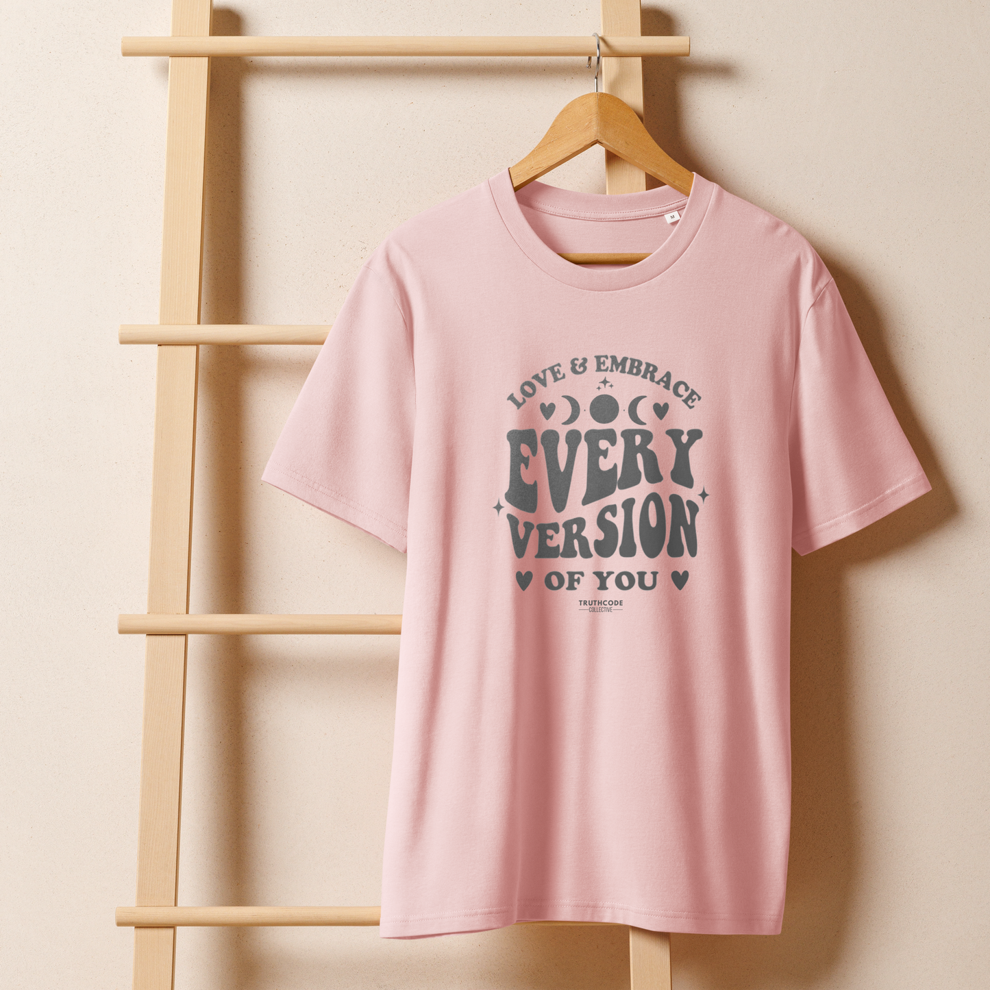 Pink organic t-shirt with motivational text 'Love & embrace every version of you' on a wooden hanger with a beige wall background