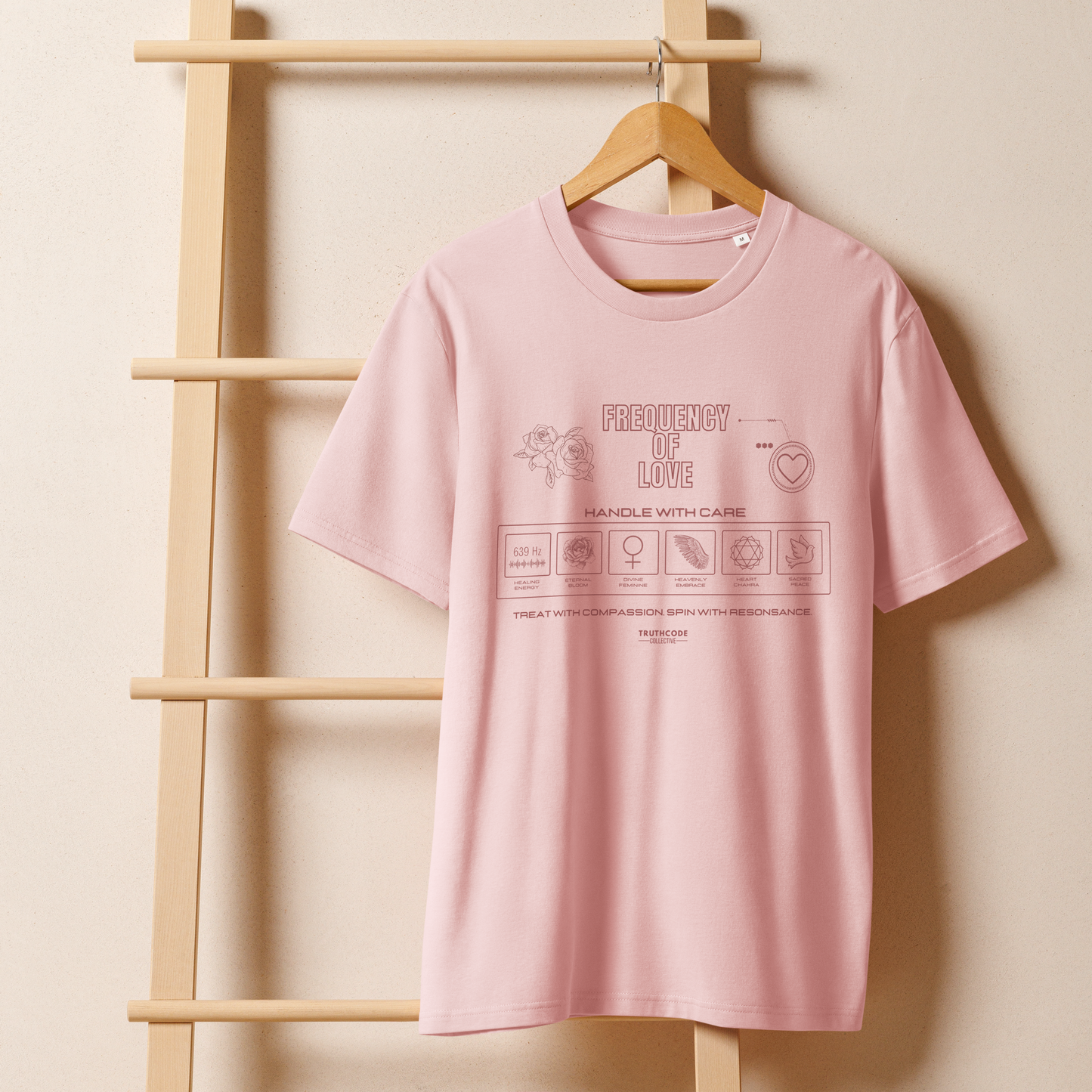 Pink organic t-shirt with text 'Frequency of love' with symbols and graphics relating to it on a wooden hanger with a beige wall background