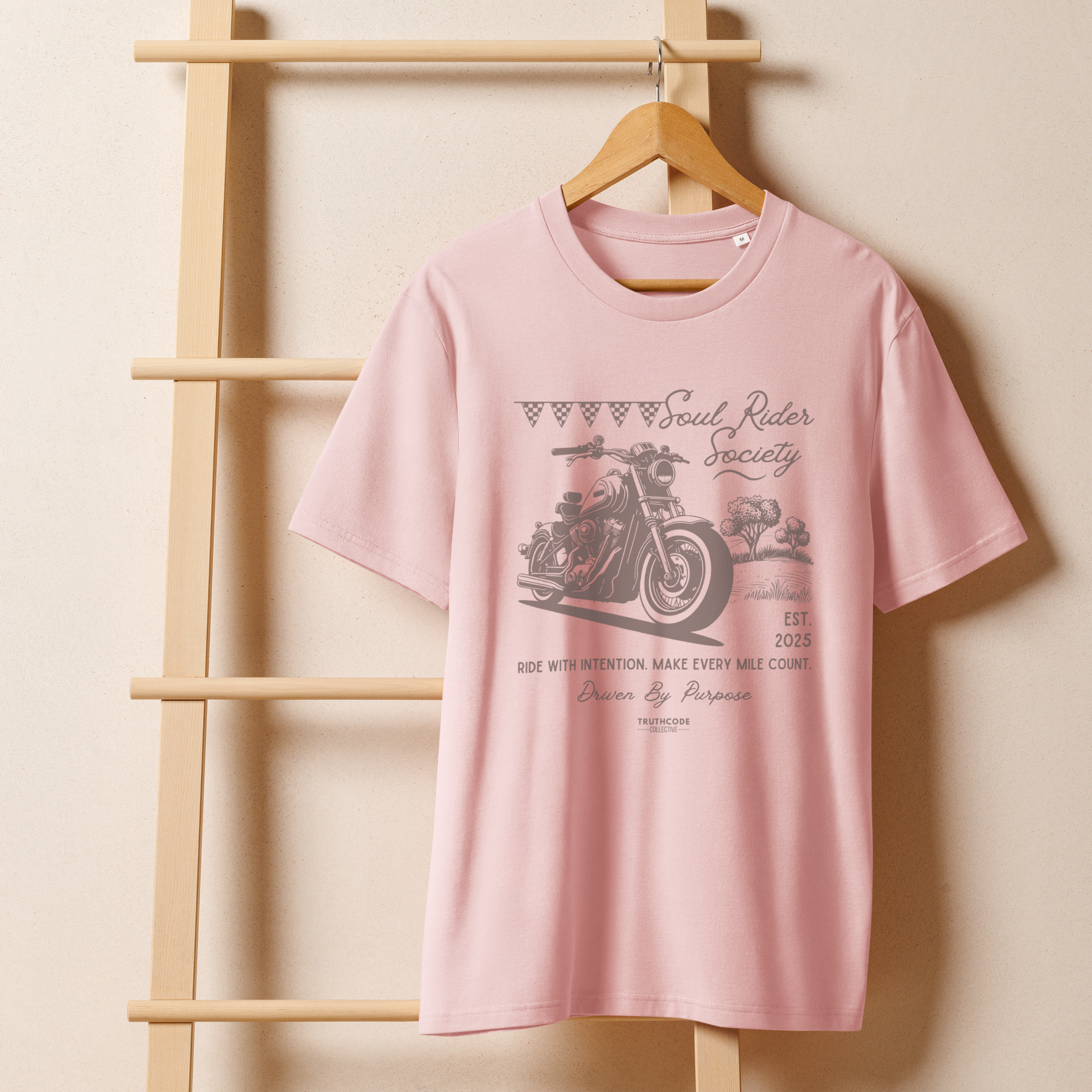 Pink organic t-shirt with motorcycle graphic and text 'Soul rider society, ride with intention, make every mile count' on a wooden hanger with a beige wall background
