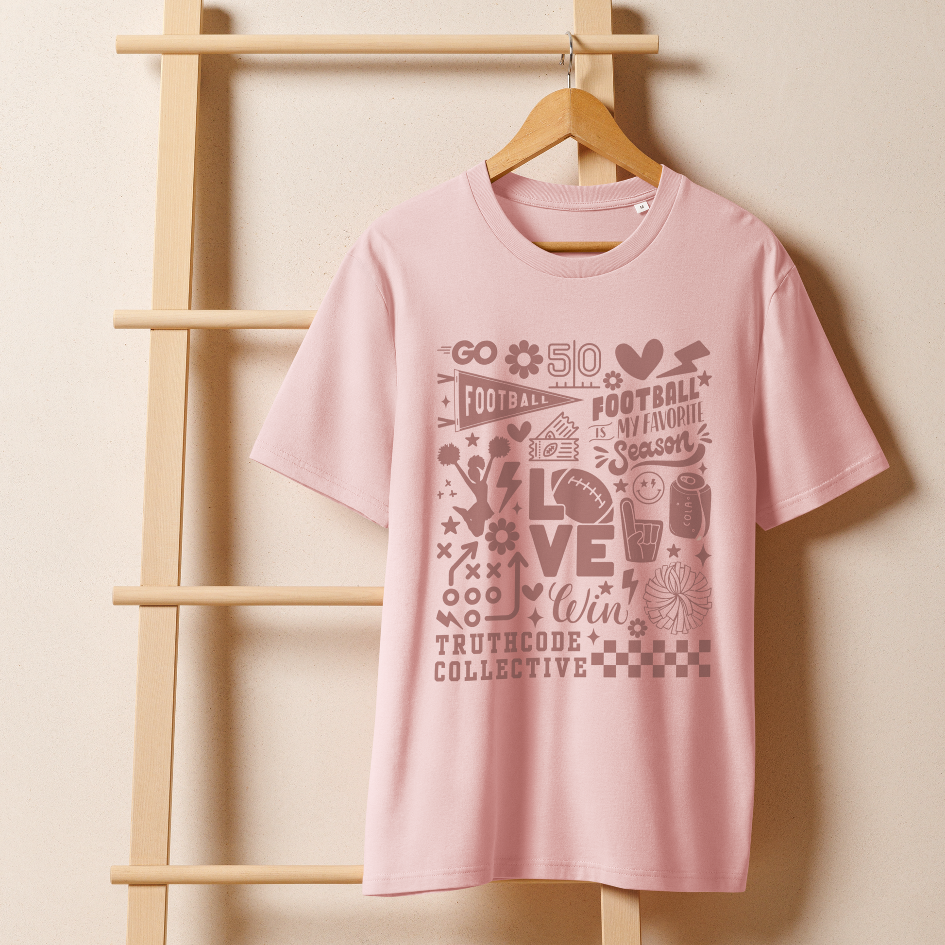 Pink organic t- shirt with football themed graphics and text 'Football is my favorite season' on a wooden hanger with a beige wall background