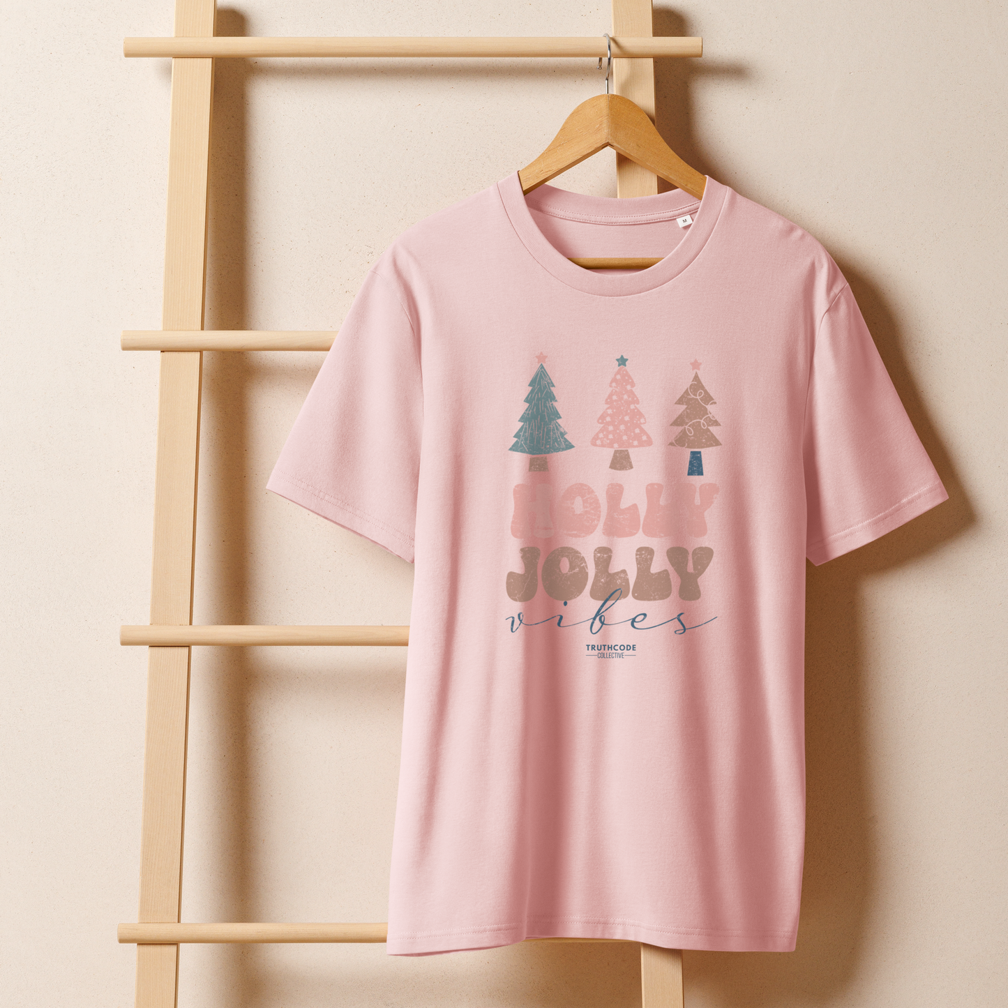 Pink organic t-shirt with Christmas tree design and 'Holly Jolly vibes' text on a wooden hanger with a beige wall backfground