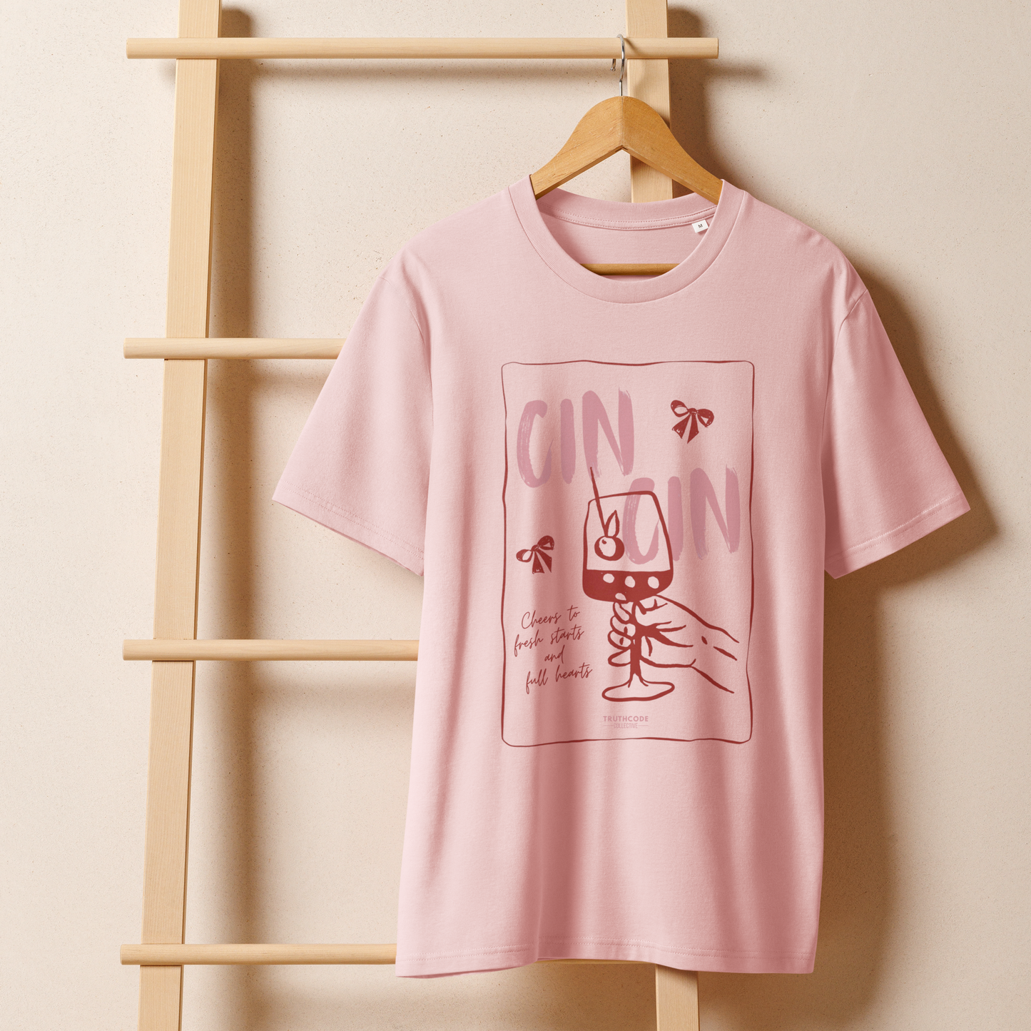 Pink organic t-shirt with text 'Cin Cin, cheers to fresh starts and full hearts' graphic design  of a glass, on a hanger with a beige wall background