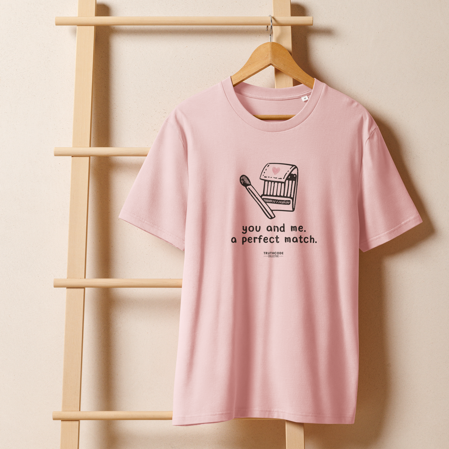 Pink organic t-shirt with match book graphic design and text 'You and me, a perfect match' on a hanger with a beige wall background