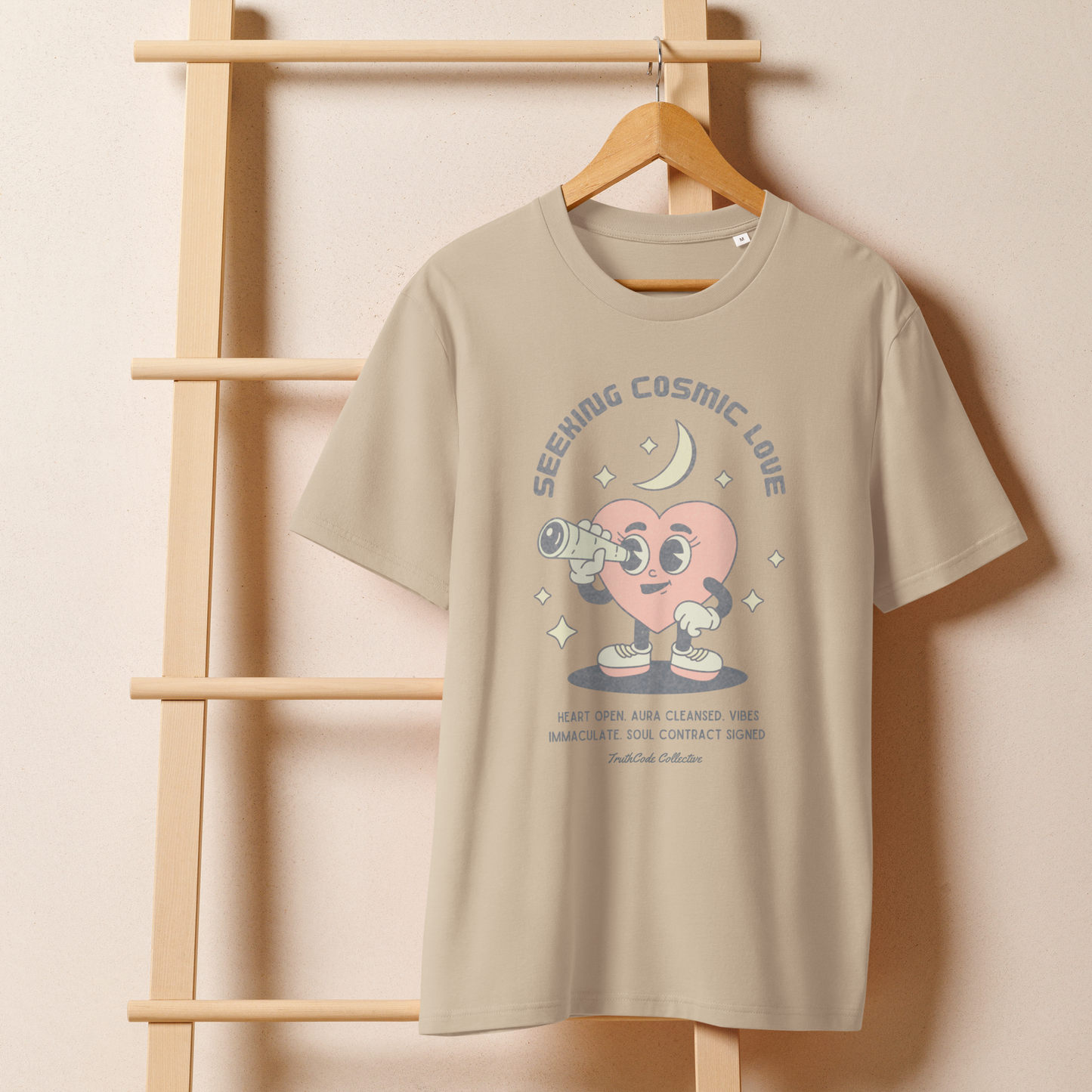 Beige organic t-shirt with a heart-shaped character holding a telescope and text 'Seeking Cosmic Love' on a wooden hanger with a beige wall background