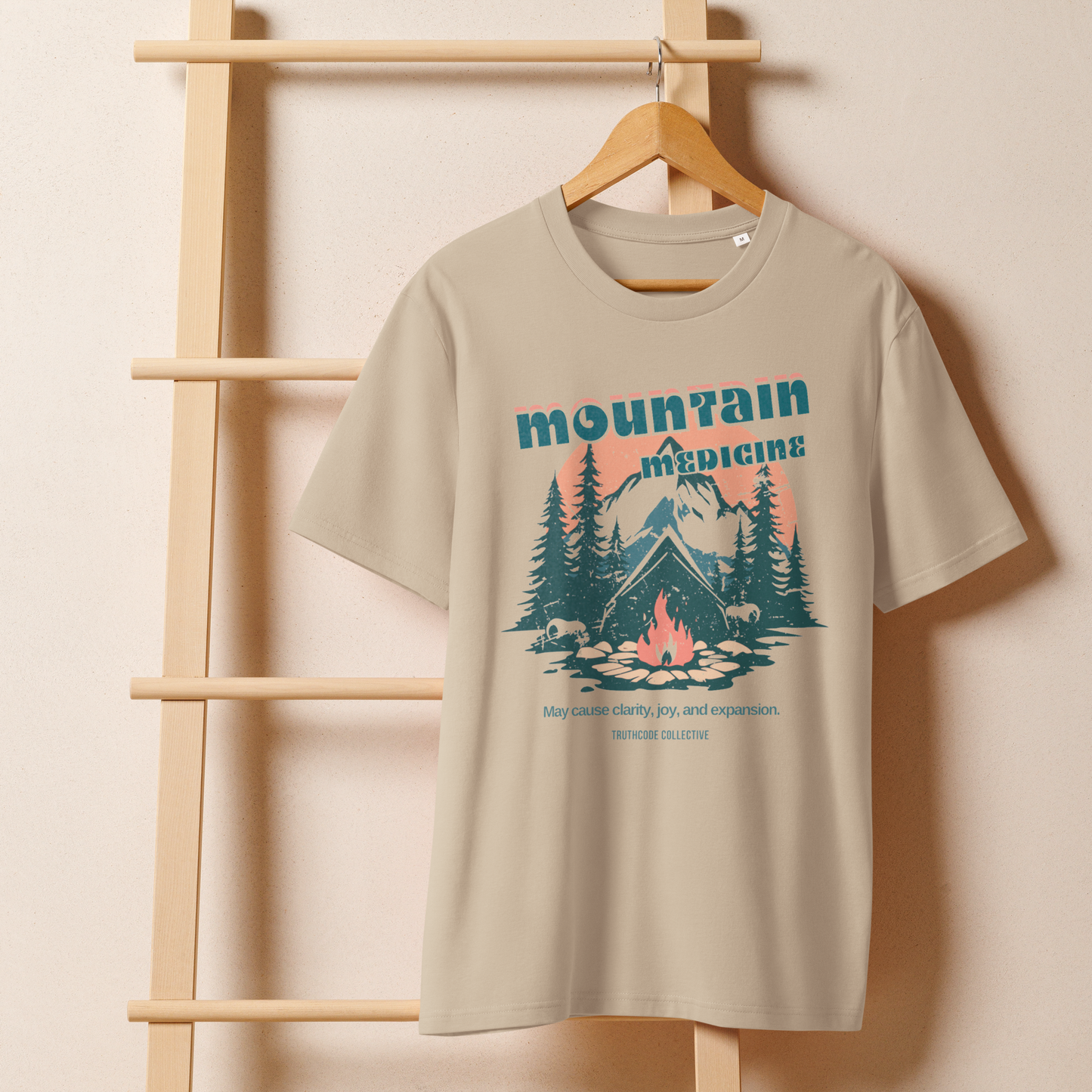 Beige organic t-shirt with mountain and campfire graphic and 'Mountain Medicine' text on a wooden hanger with a beige wall background