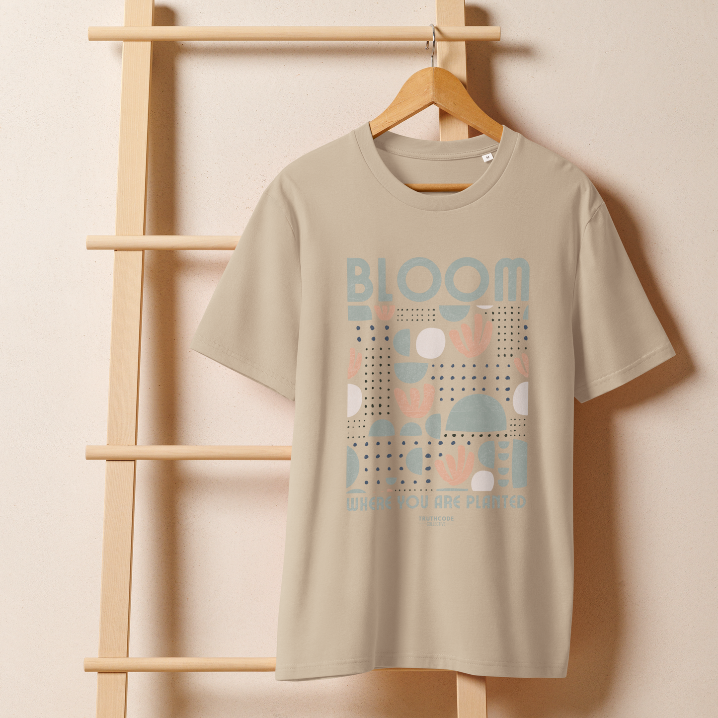 Beige organic t-shirt with 'Bloom Where You Are Planted' design on a wooden hanger with a beige wall background