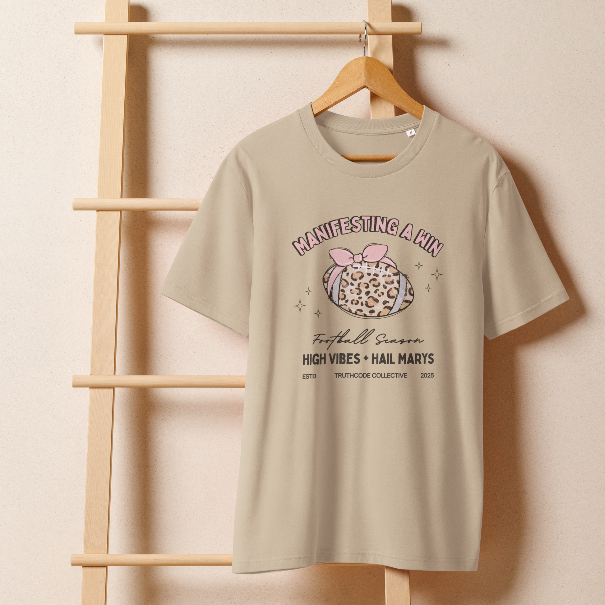 Beige organic t-shirt with girly football graphic and text 'Manifesting a win' and 'Football season, high vibes and hail marys' on a wooden hanger with a beige wall background