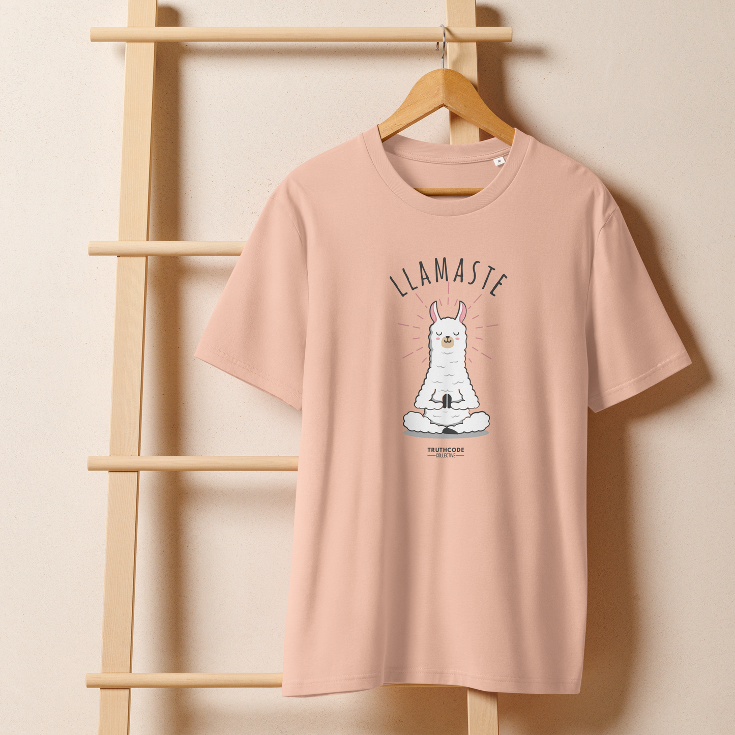 Peach organic t-shirt with a llama graphic in meditative pose and 'LLAMASTE' text on a wooden hanger with a beige wall background