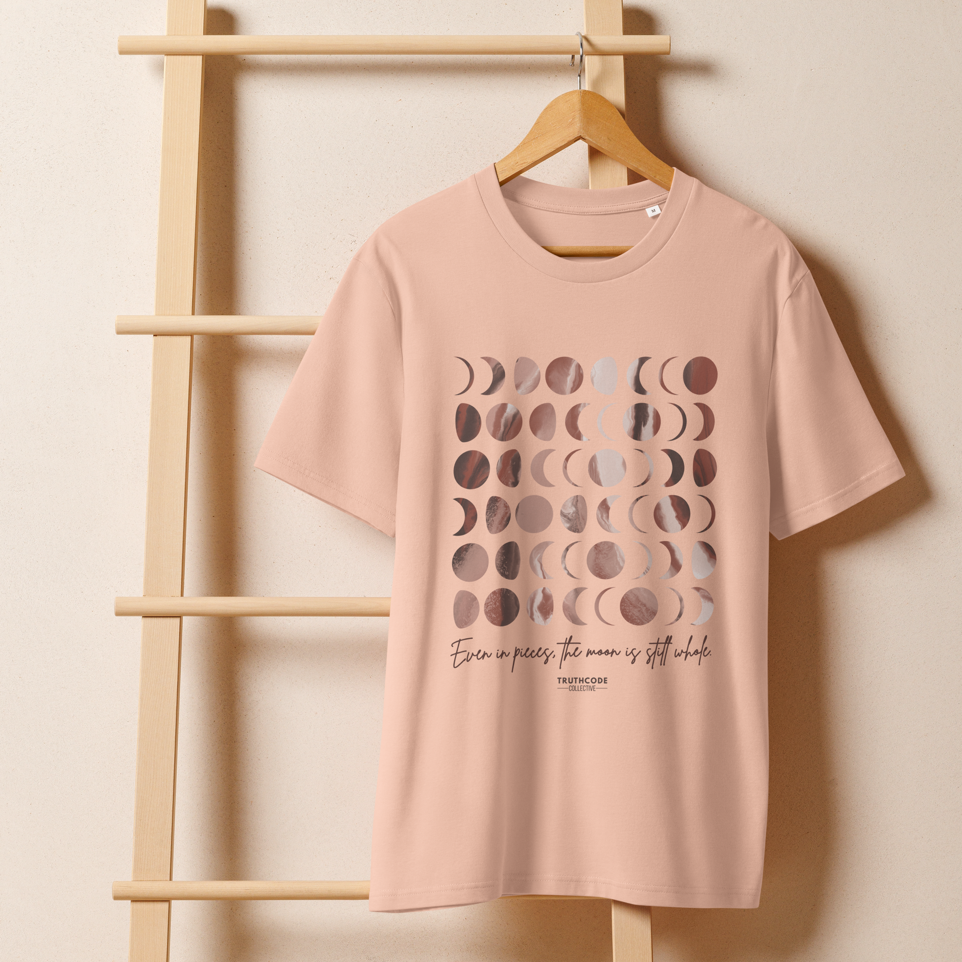Peach organic t-shirt with rose gold moon phase design and text 'Even in pieces, the moon is still whole' on a wooden hanger with a beige wall background
