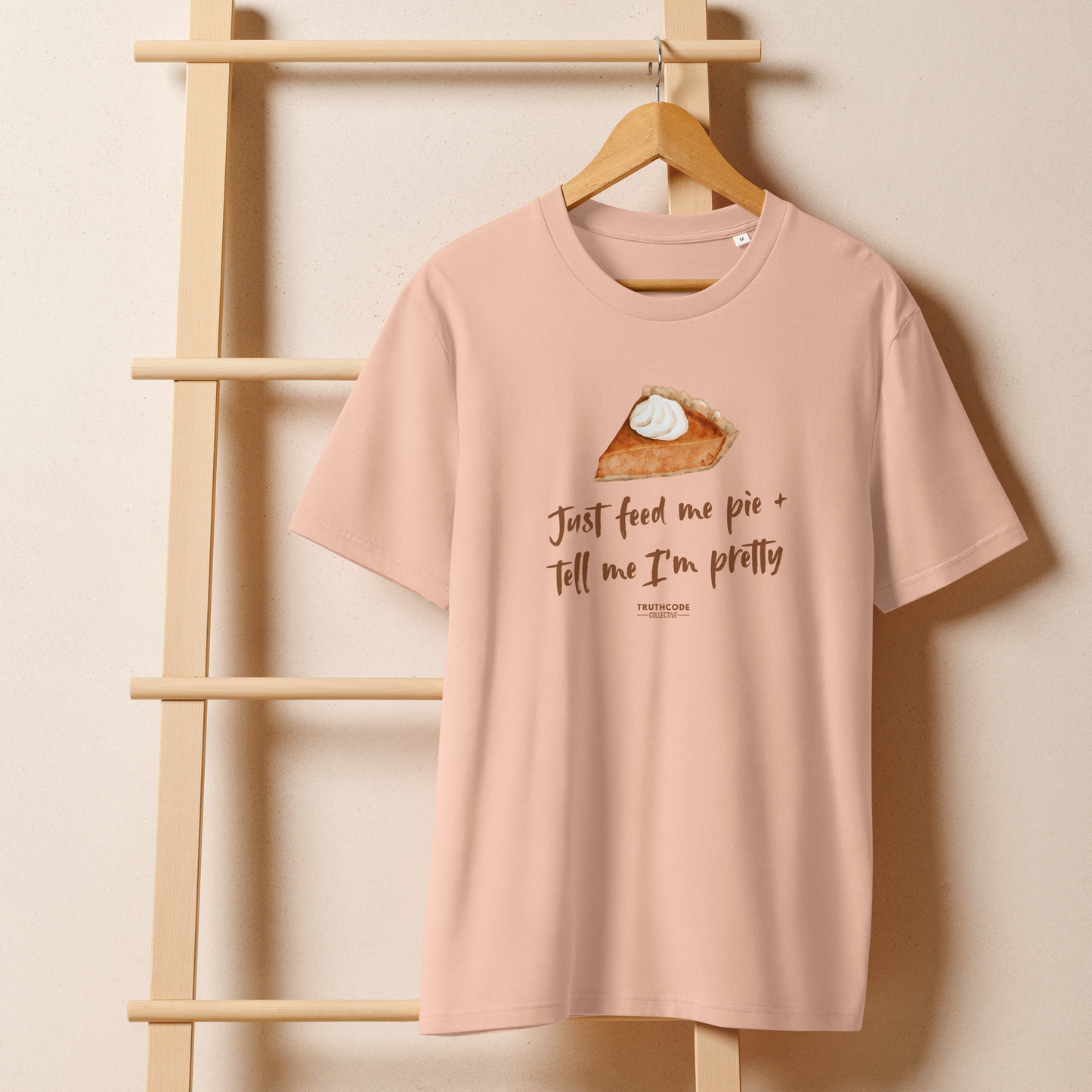 Peach organic t-shirt with a pie graphic and text 'Just feed me pie + tell me I'm pretty' on a wooden hanger with a beige wall background
