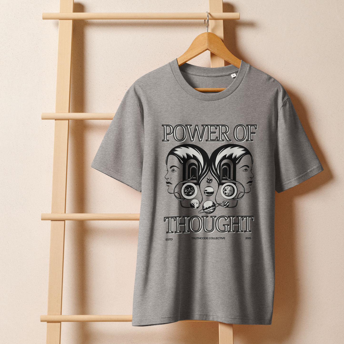 Gray organic t-shirt with 'Power of Thought' text and design of graphics of the universe coming from 2 heads on a wooden hanger with a beige wall in the background