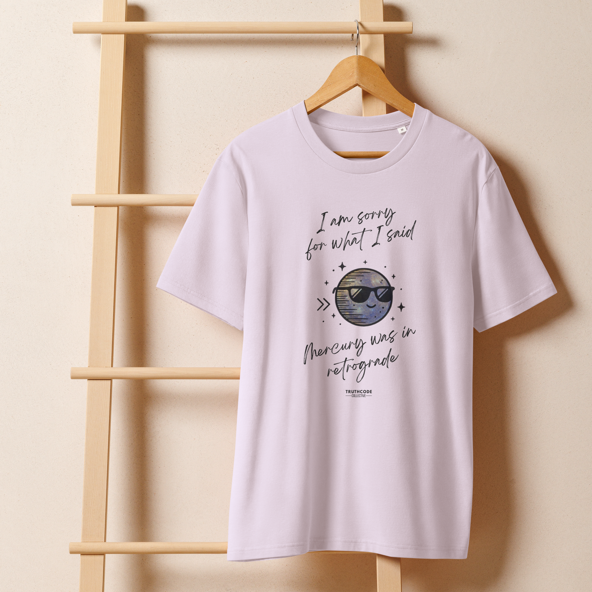 Lavender organic t-shirt with graphic of a planet wearing sunglasses design and text reading 'I'm sorry for what i said, Mercury was in retrograde' on a wooden hanger with a beige wall in the background