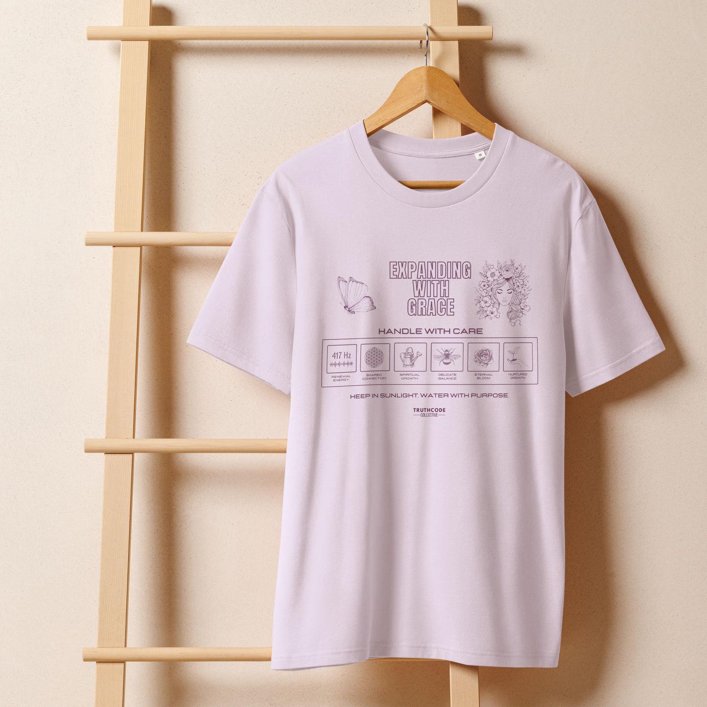 Lavender organic t-shirt with text 'Expanding with grace' with symbols and graphics on a wooden hanger against a beige wall.