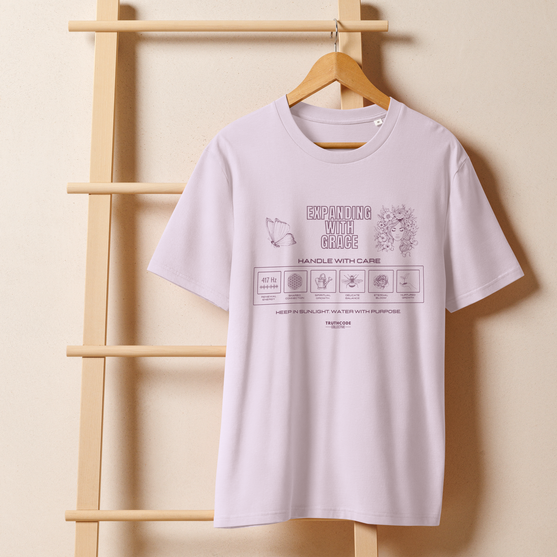 Lavender organic t-shirt with text 'Expanding with grace' with symbols and graphics on a wooden hanger against a beige wall.