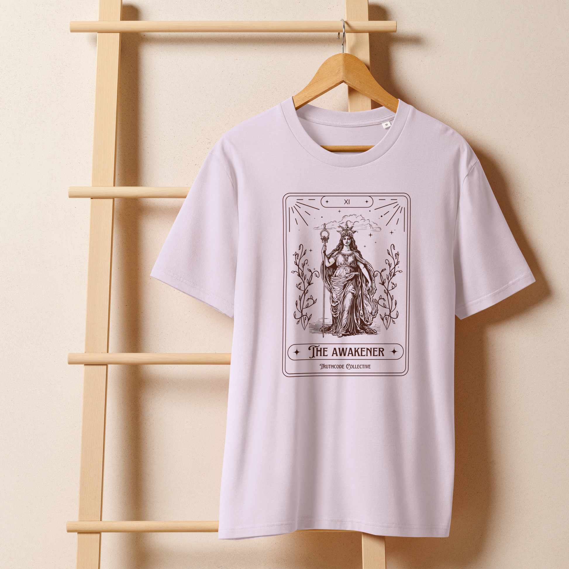 Lavender organic t-shirt with 'The Awakener' tarot card design on a wooden hanger with beige wall background