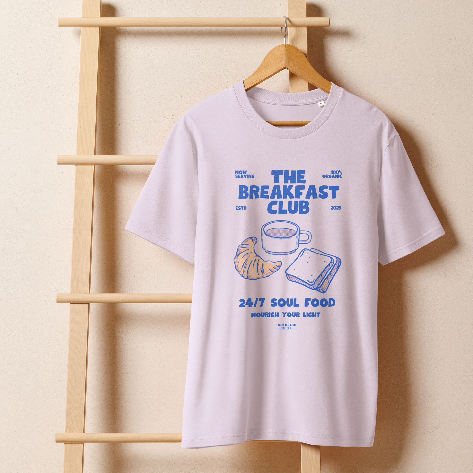 Lavender organic t-shirt with 'The Breakfast Club' graphic design featuring a coffee cup, croissant, and sandwich on a hanger with a beige wall background