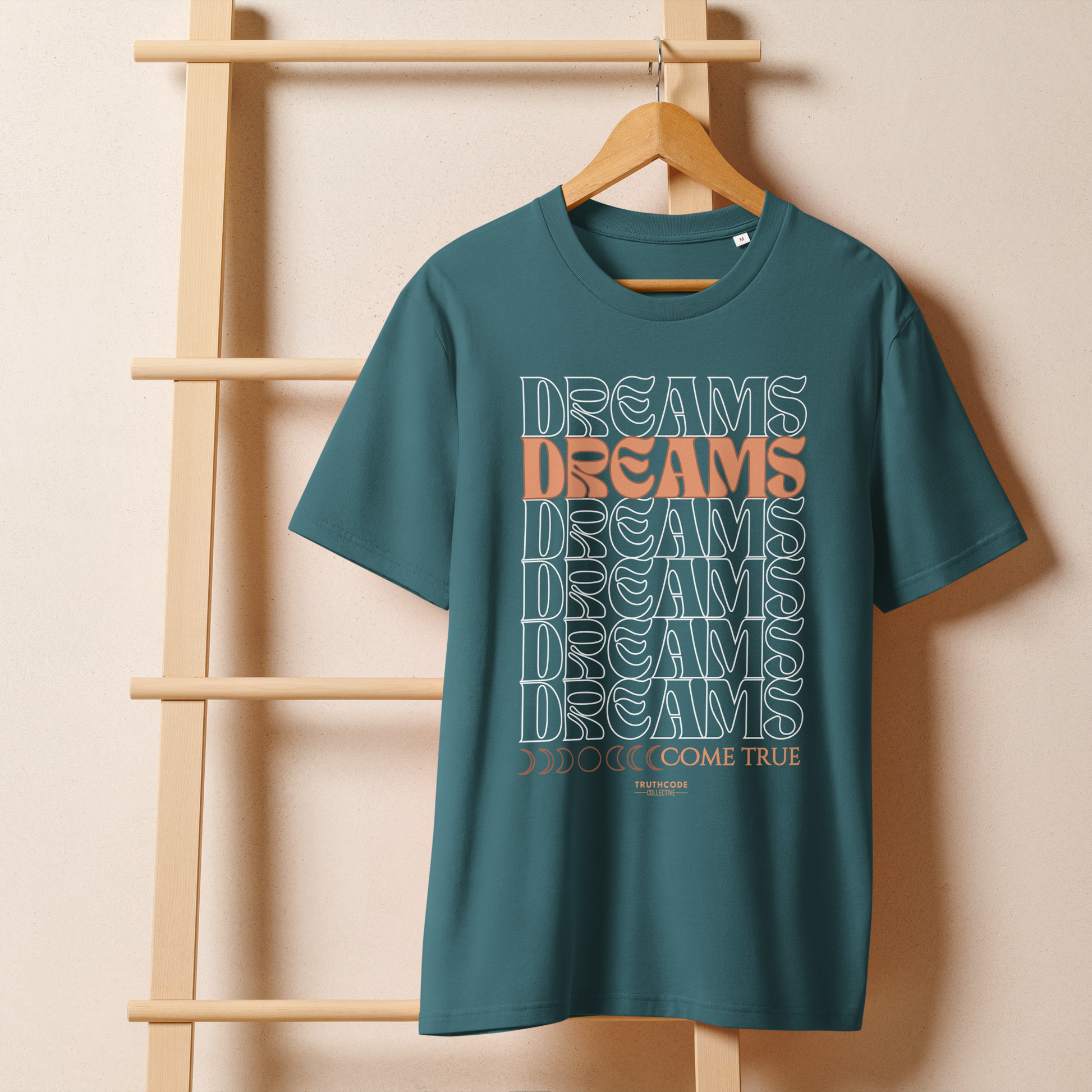 Teal organic t-shirt with 'Dreams Come True' text on a wooden hanger against a beige wall.