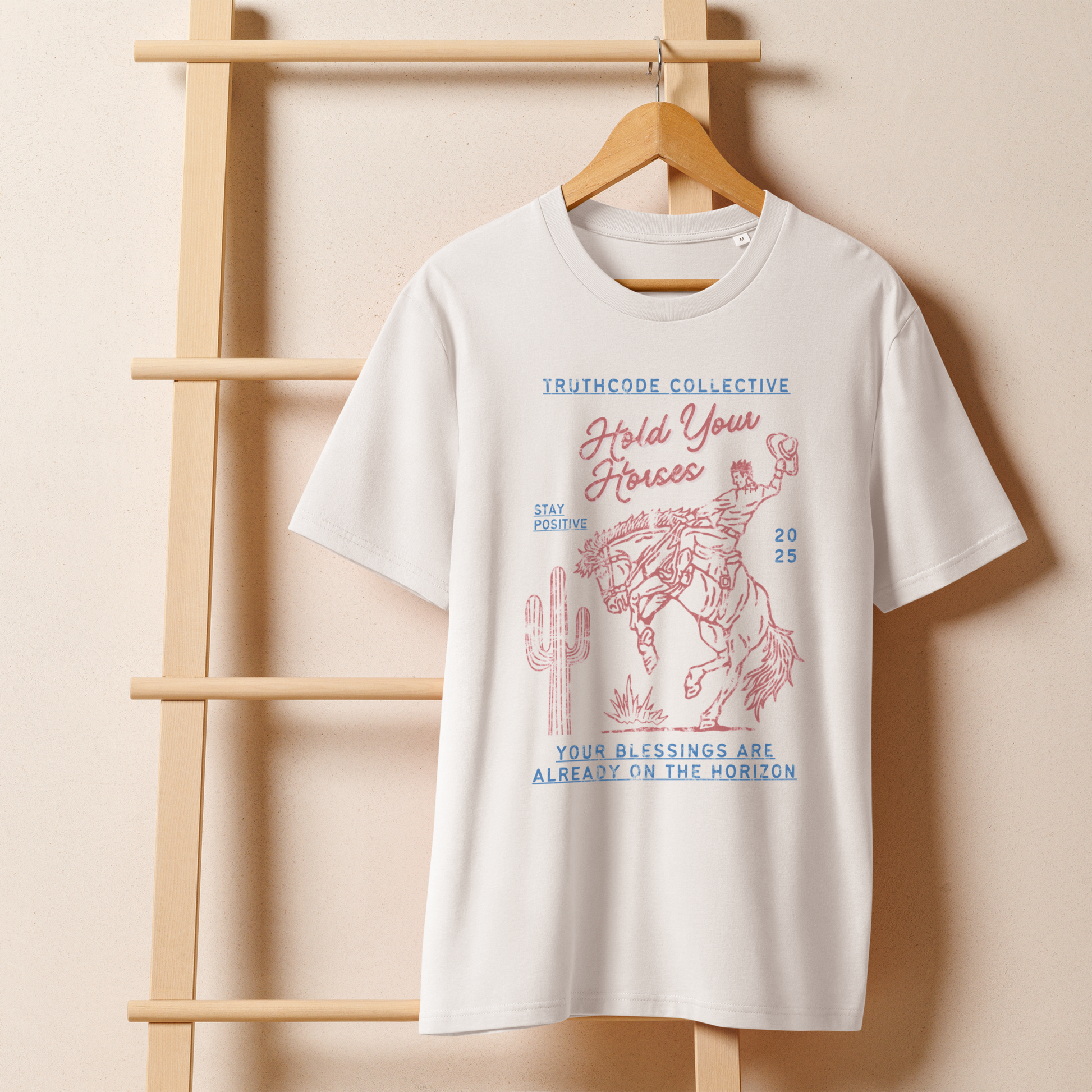 White organic t- shirt with a western style graphic of a cowboy on a bucking bronco, with the text 'Hold your horses, your blessings are already on the horizon' hanging on a wooden hanger