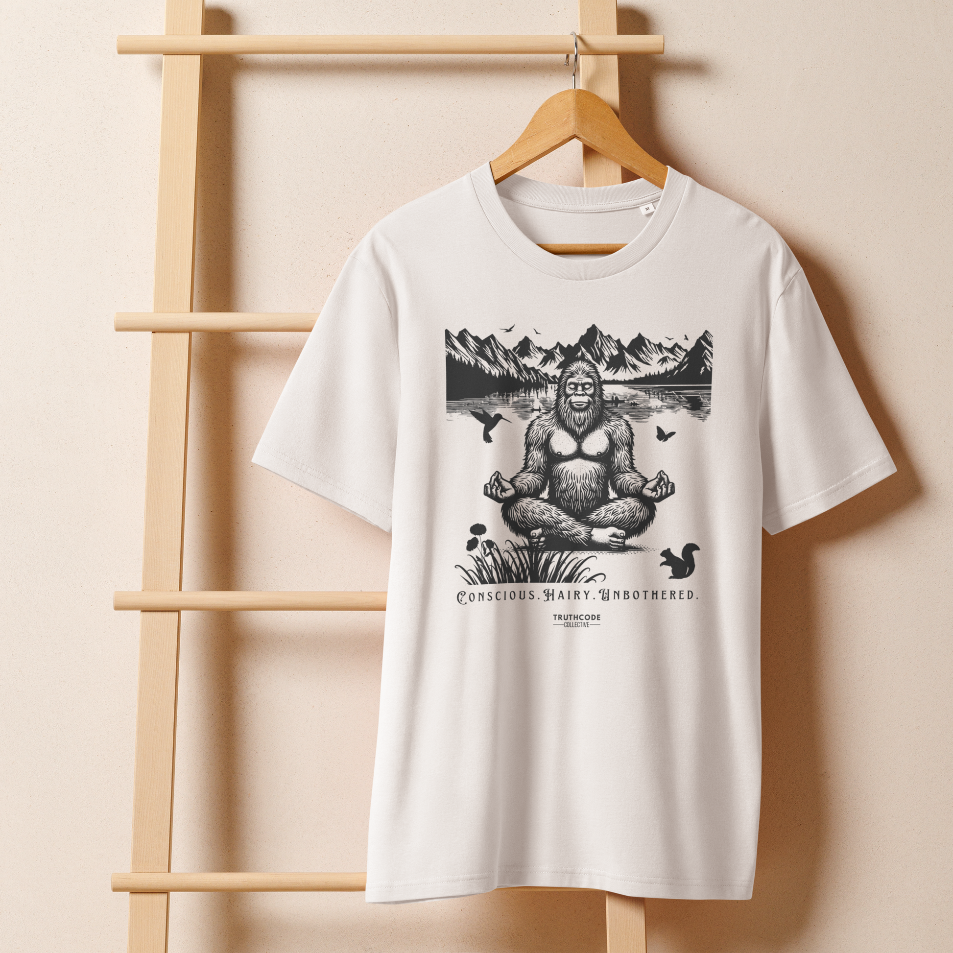 White organic t-shirt with a graphic of a meditating Sasquatch in a mountain setting and text reading 'Conscious, hairy, and unbothered' hanging on a wooden hanger