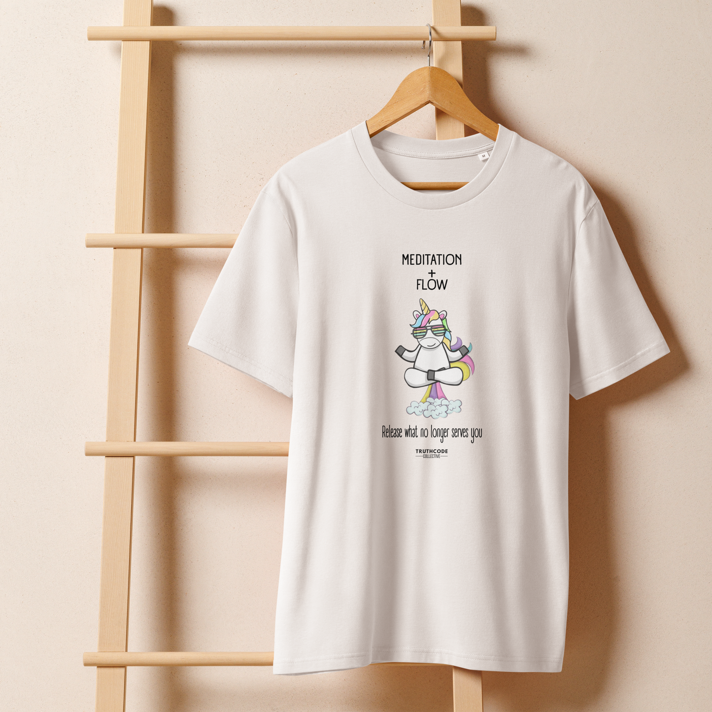 White organic t-shirt with a humorous farting unicorn graphic and text reading 'Meditation + Flow, release what no longer serves you' on a wooden hanger with a beige wall in the background