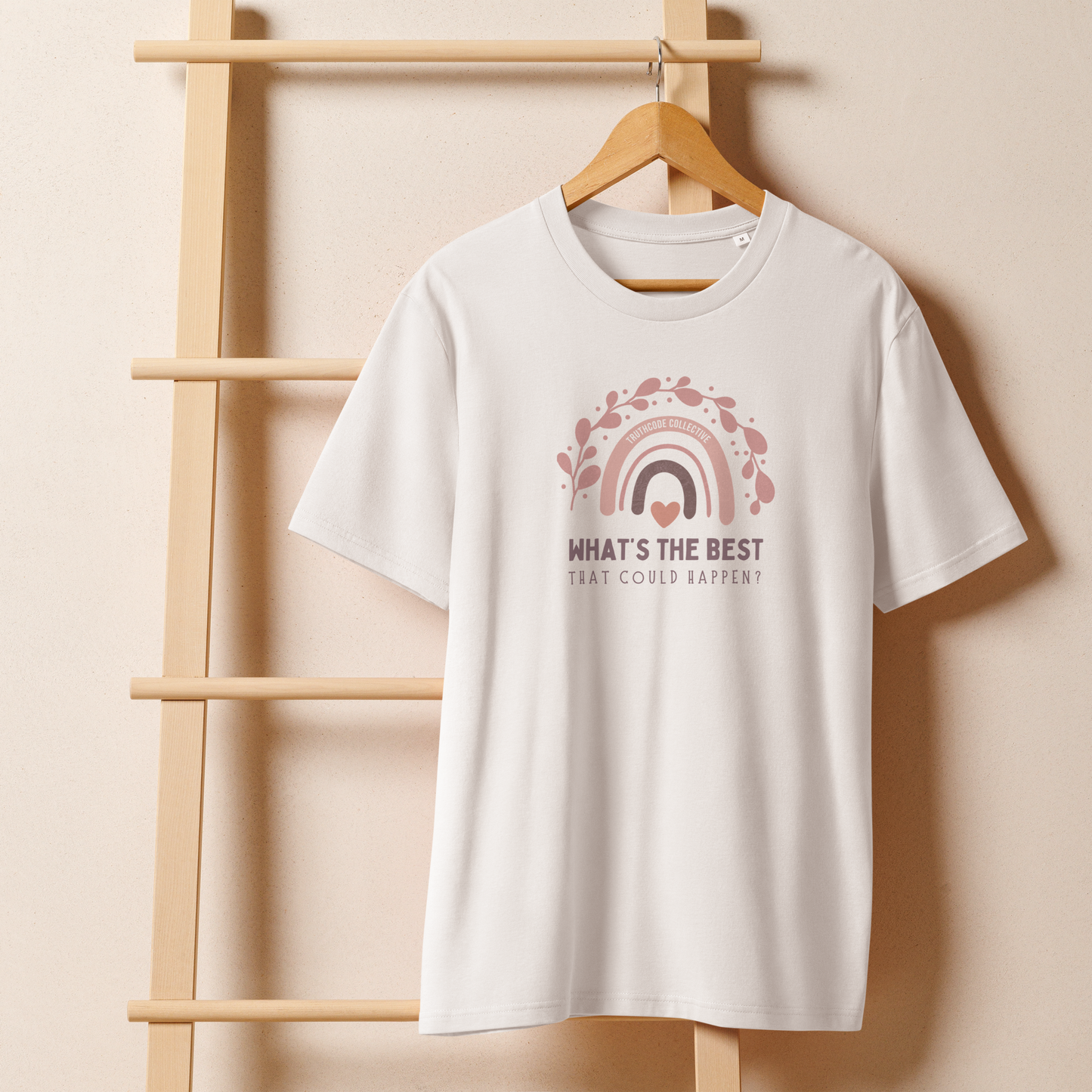 White organic t-shirt with a pink rainbow and text 'What's the best that could happen?' design on a wooden hanger with a beige wall background