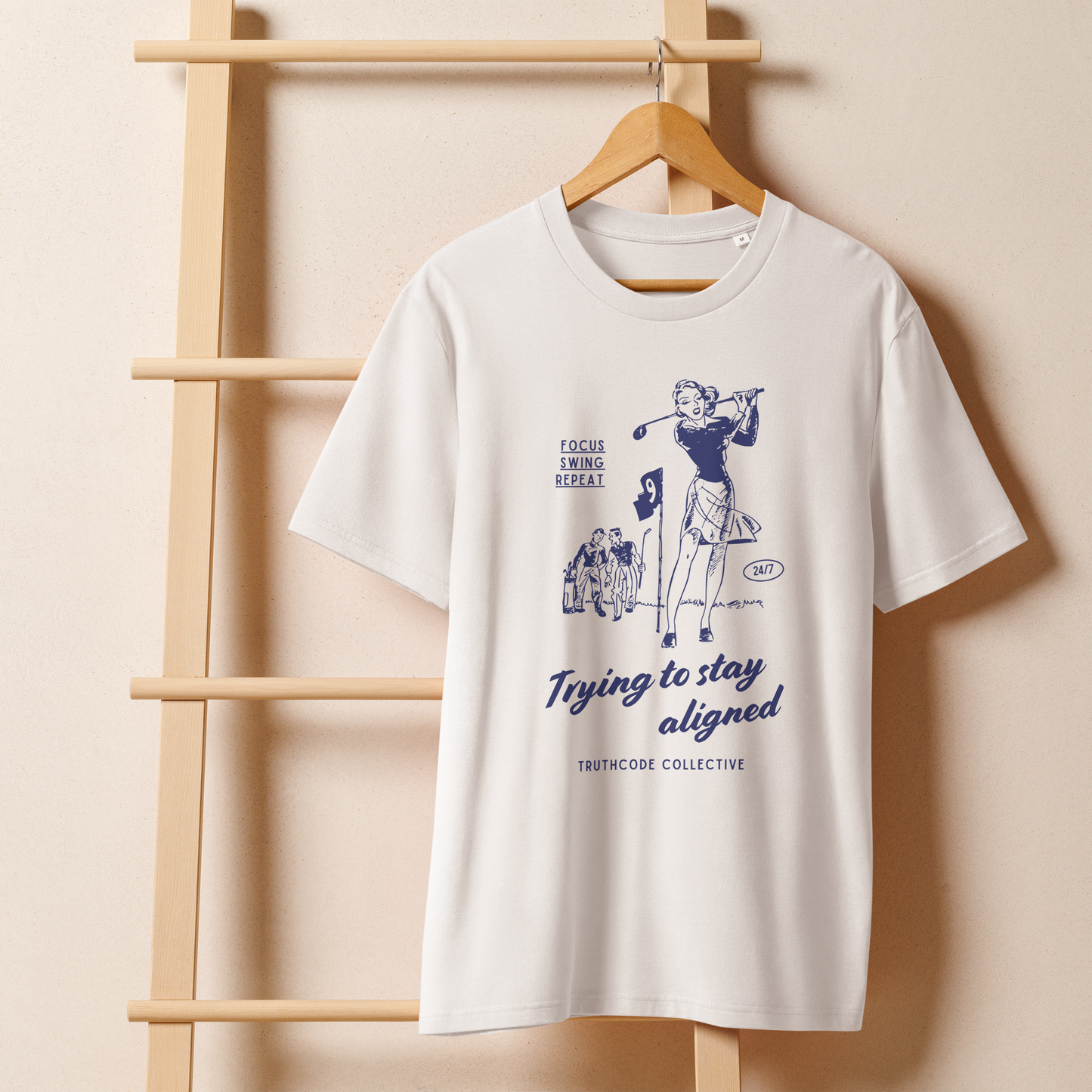 White organic t-shirt with blue vintage graphic of a woman golfing and text 'Trying to stay aligned' and 'Focus, swing, repeat' on a wooden hanger with beige wall background