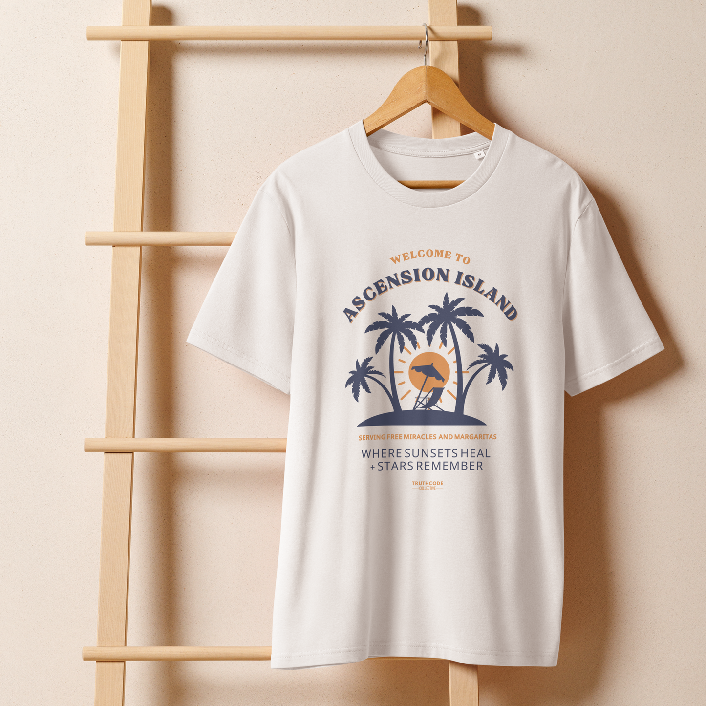 White organic t-shirt with palm tree design and text 'Ascension island, where sunsets heal + stars remember' on a wooden hanger with a beige wall background