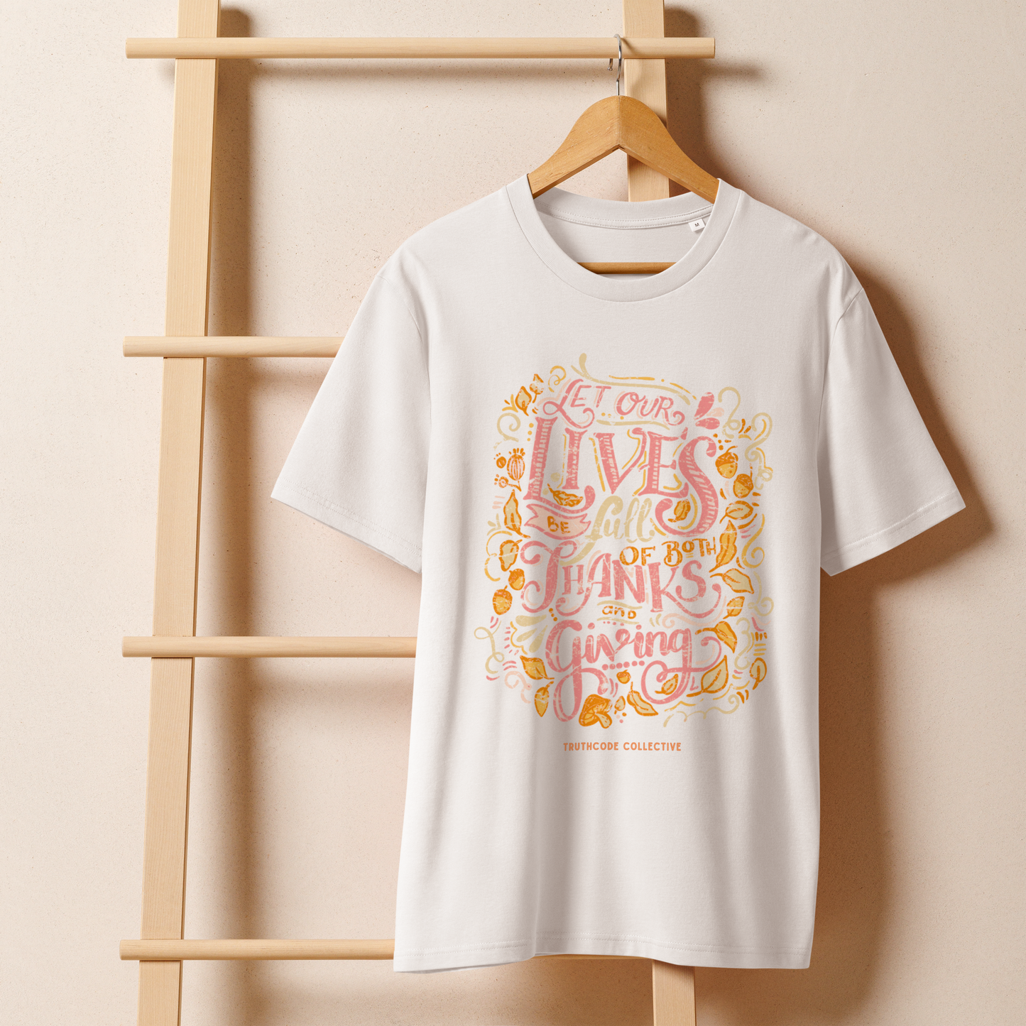 White organic t-shirt with colorful text design 'Let our lives be full of both thanks and giving' on a wooden hanger with a beige wall background