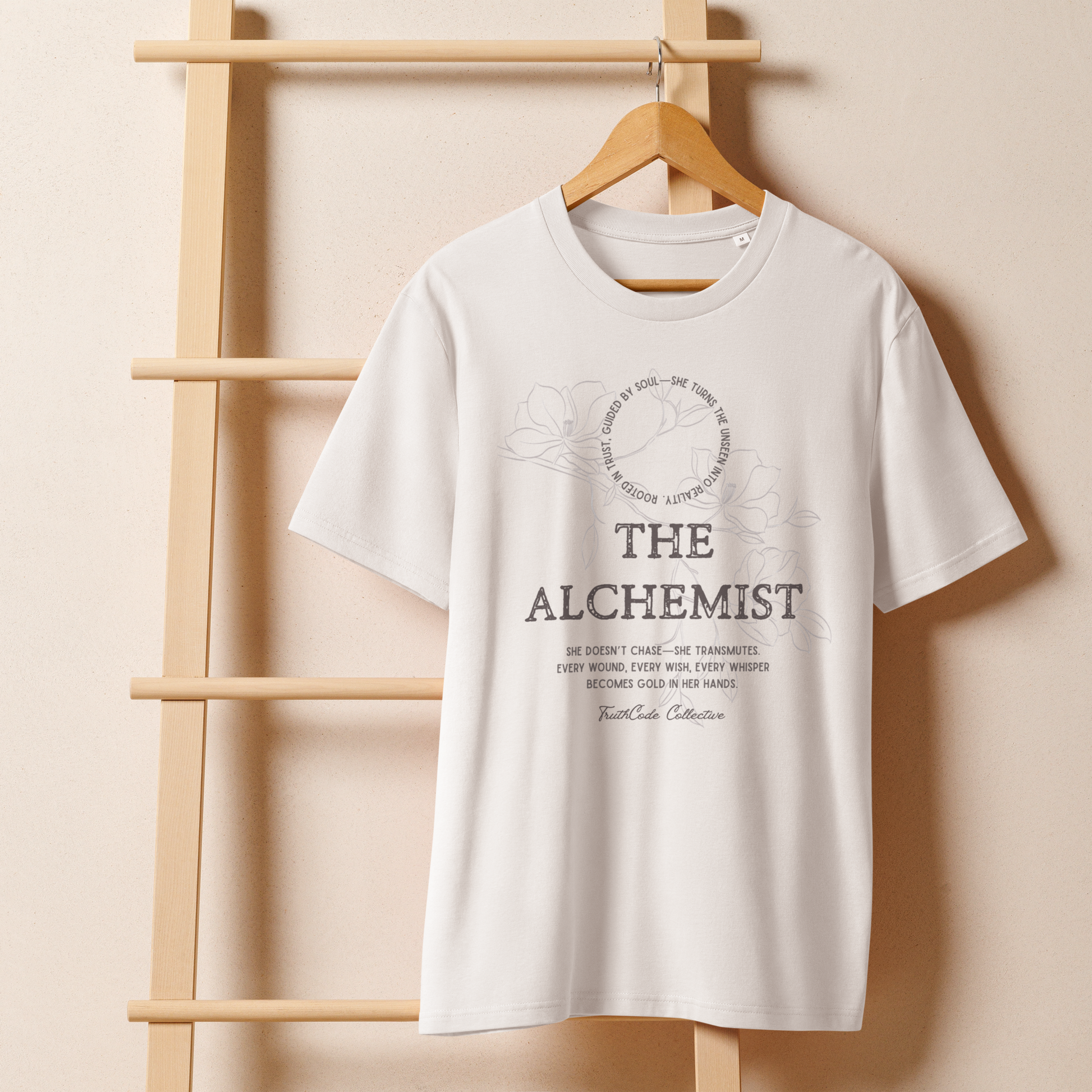 White organic t-shirt with text 'The Alchemist, she doesn't chase- she transmutes...' and subtle floral background design on a wooden hanger with a beige wall background