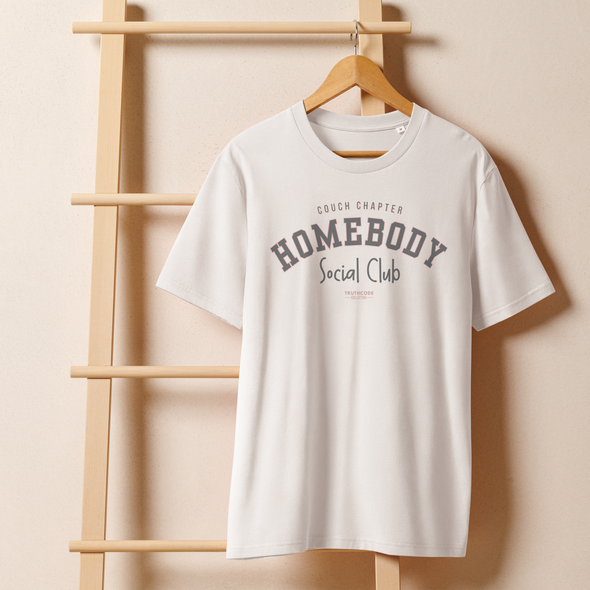 White organic t-shirt with 'Homebody Social Club' text on a wooden hanger with a beige wall background