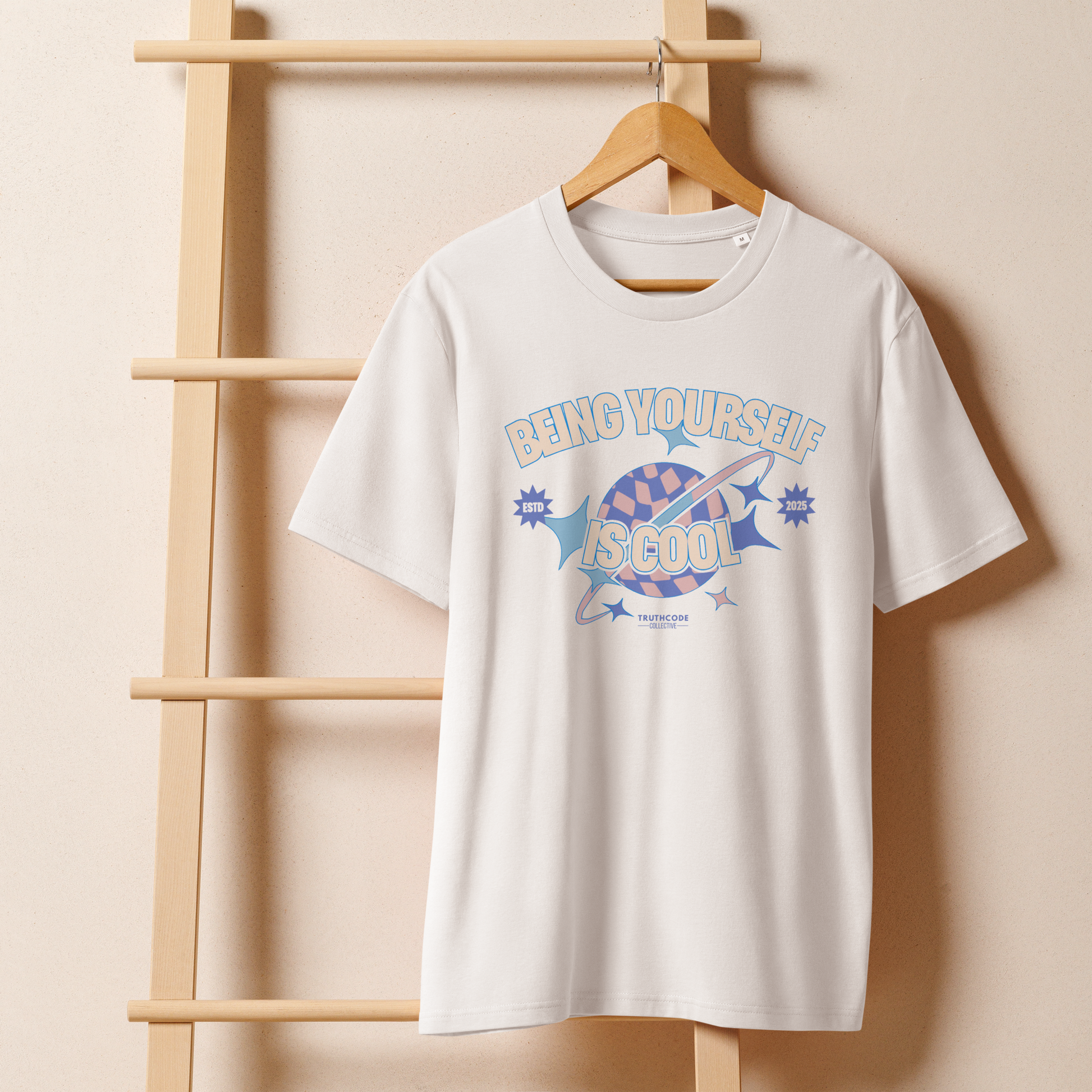 White organic t-shirt with a colorful graphic and text 'Being Yourself is Cool' on a hanger with a beige wall background