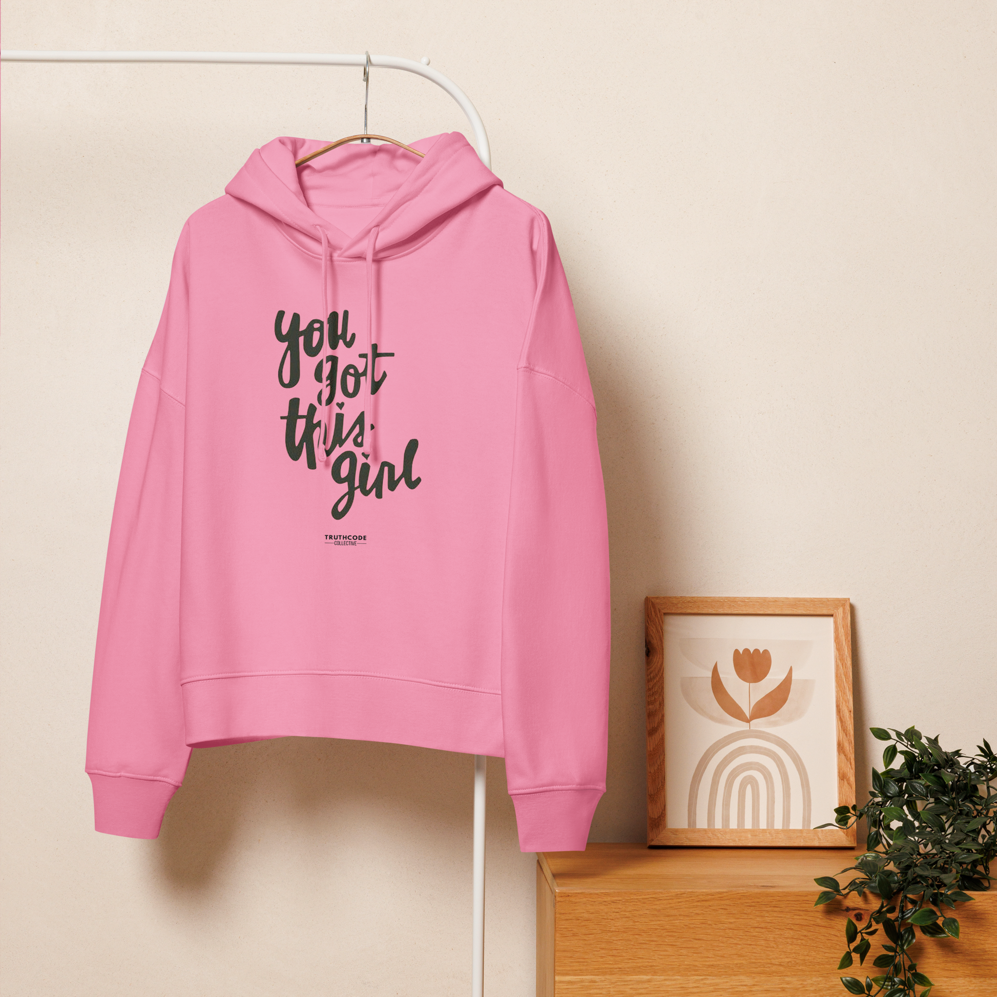 Pink organic hoodie with text 'You got this girl', on a hanger with a beige wall background