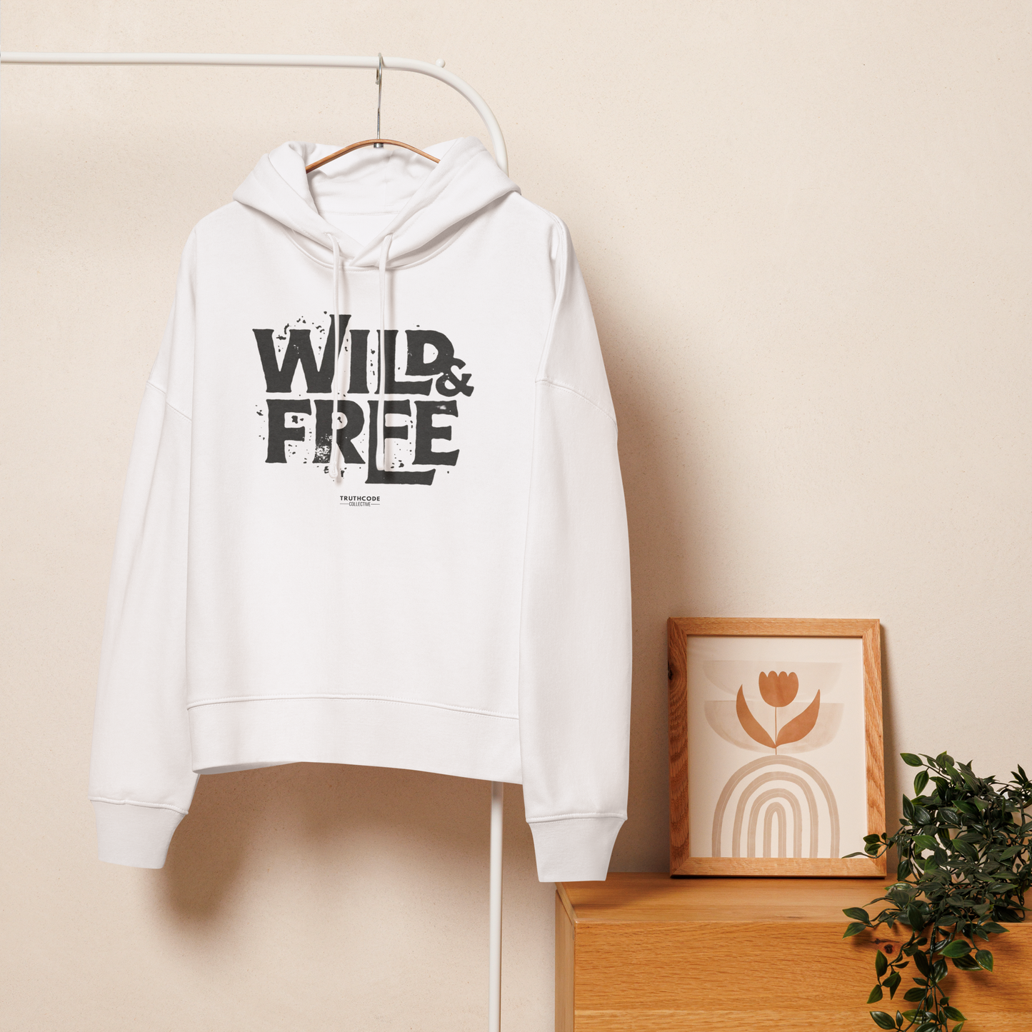 White organic hoodie with 'Wild & Free' text design, on a hanger with a beige wall background